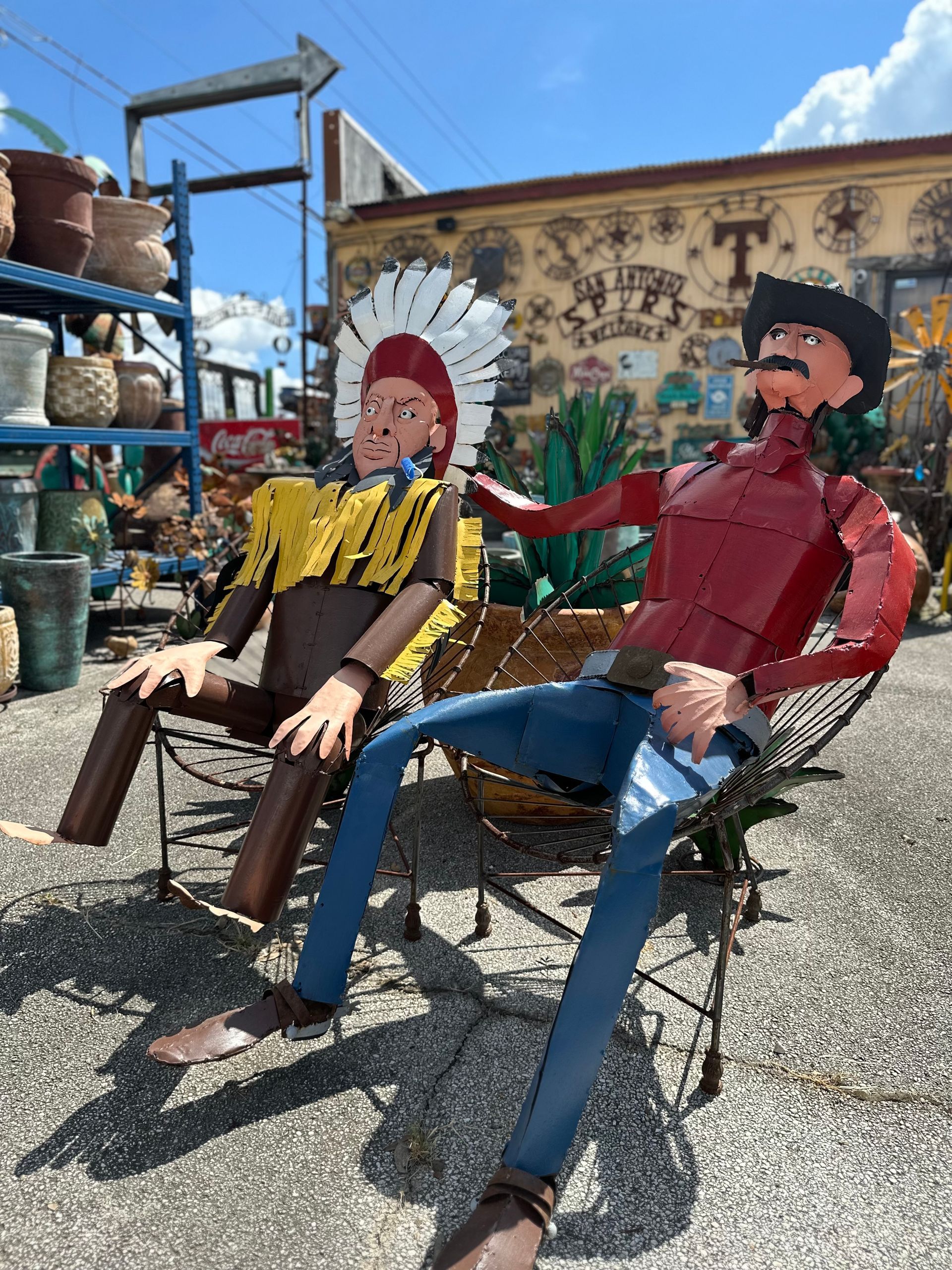 Two metal sculptures, a Native American and a cowboy, seated in chairs. They sit outdoors in front of a building with various items for sale.