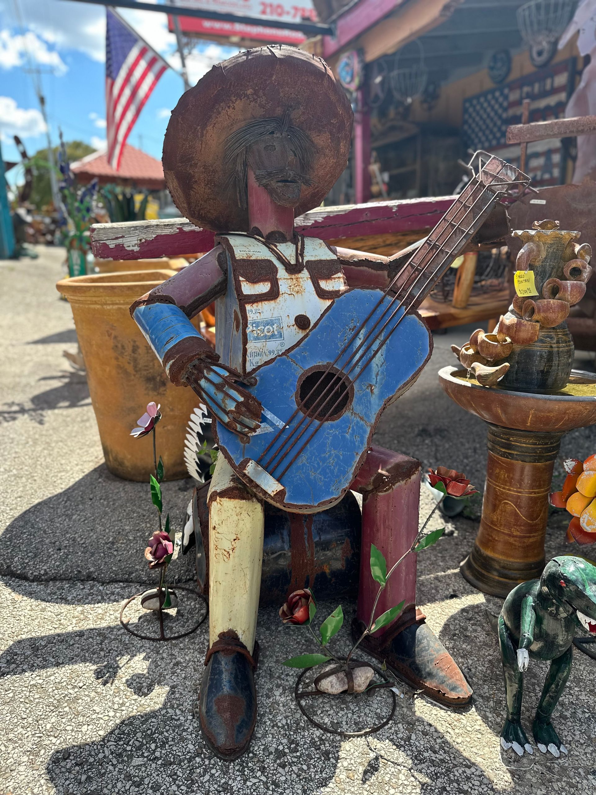 Metal sculpture of a seated mariachi playing a blue guitar, in an outdoor market with an American flag in the background.