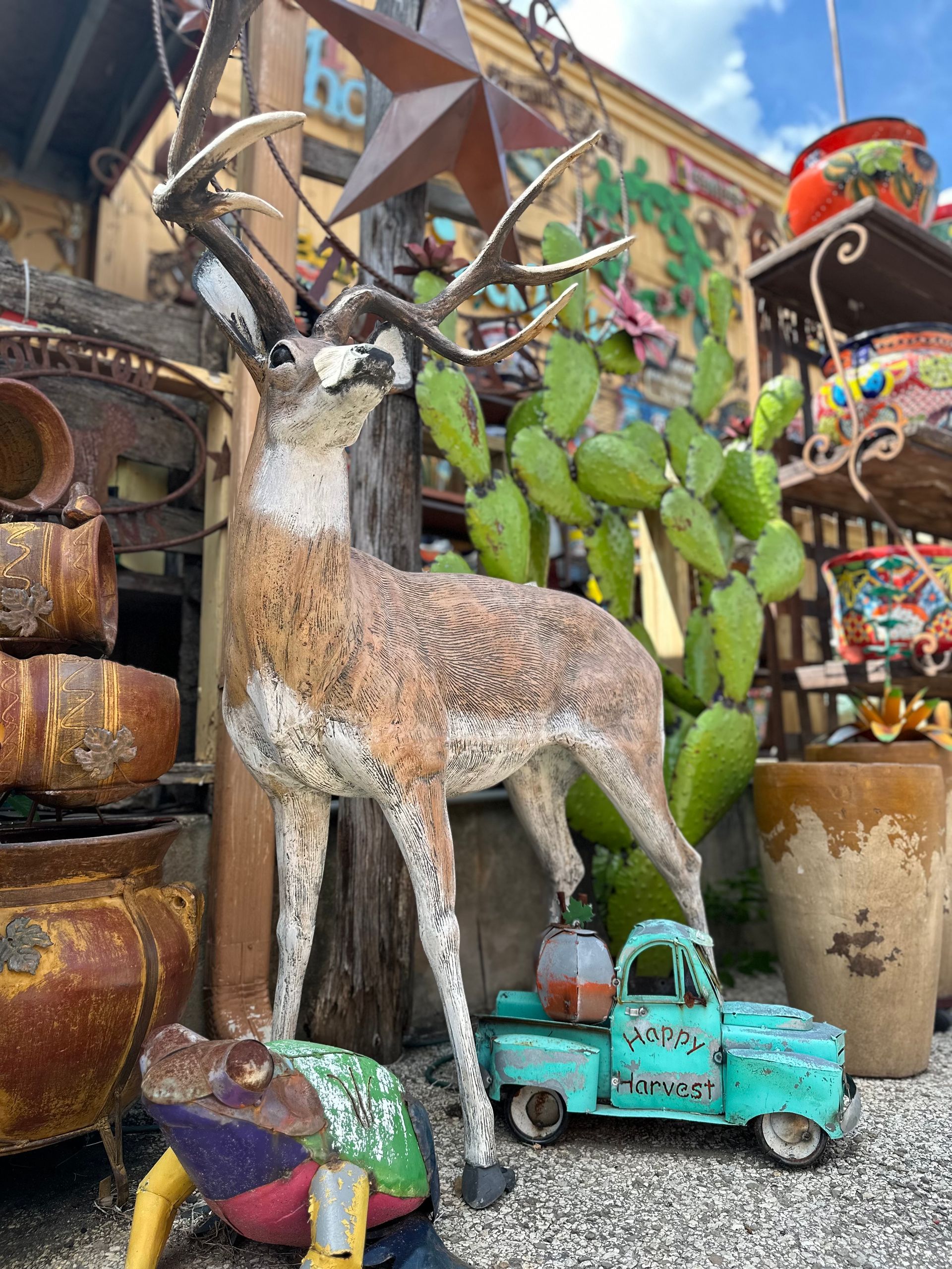 A deer statue stands in front of a cactus and a turquoise truck, surrounded by rustic decor and pottery.