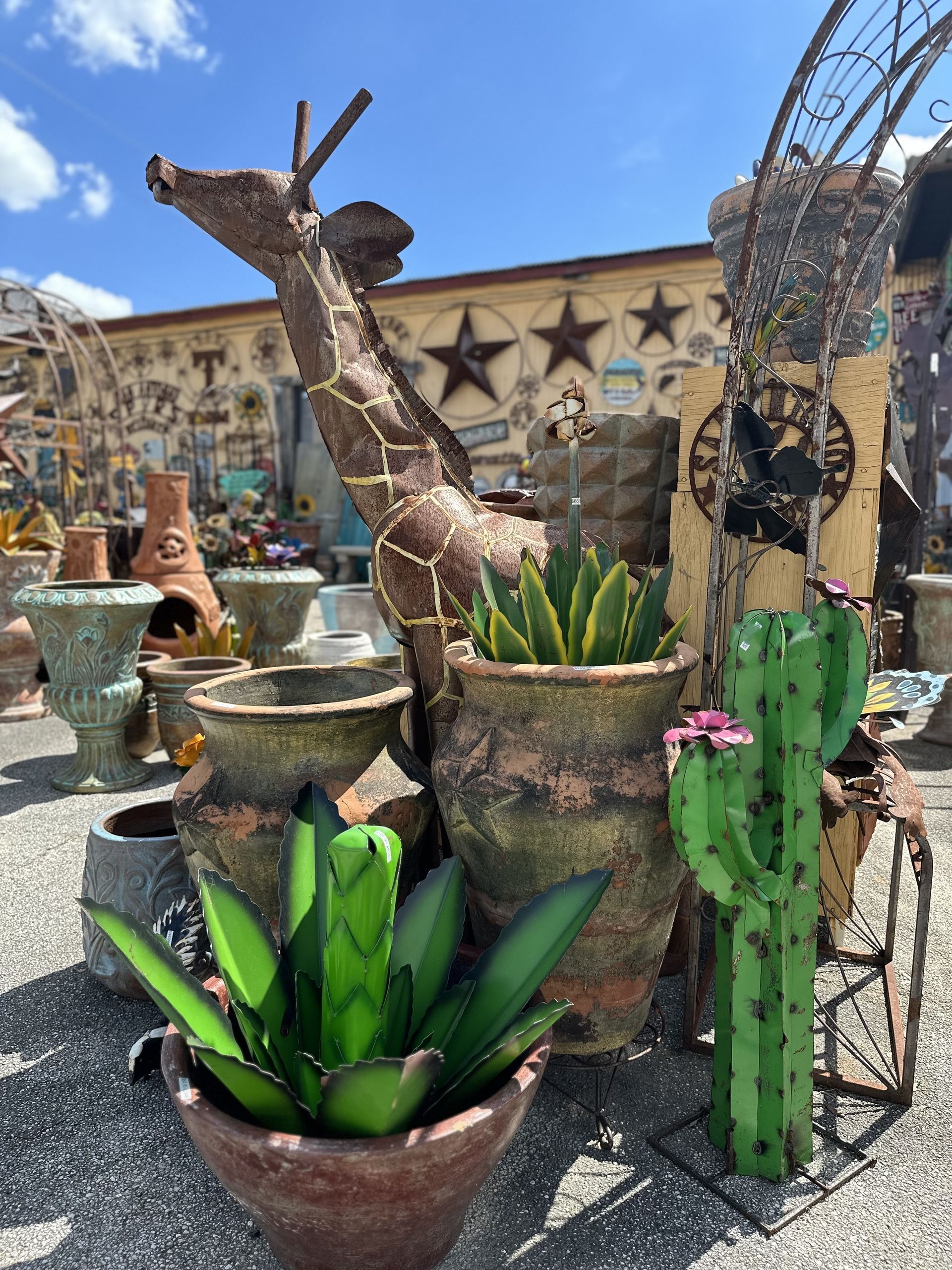 Metal giraffe sculpture and cactus with potted plants, set against a background of a building with star decorations under a blue sky.