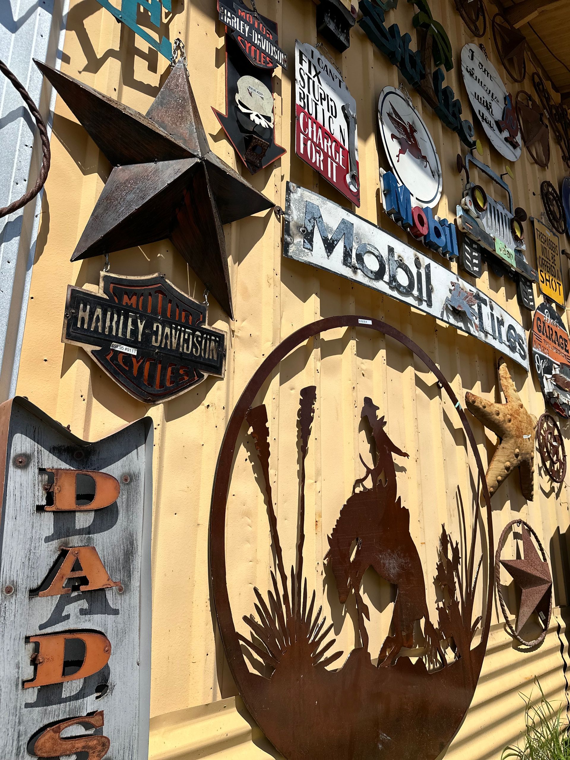 Metal signs and art mounted on a weathered, yellow-toned wall, including a star, Harley Davidson logo, and howling wolf silhouette.