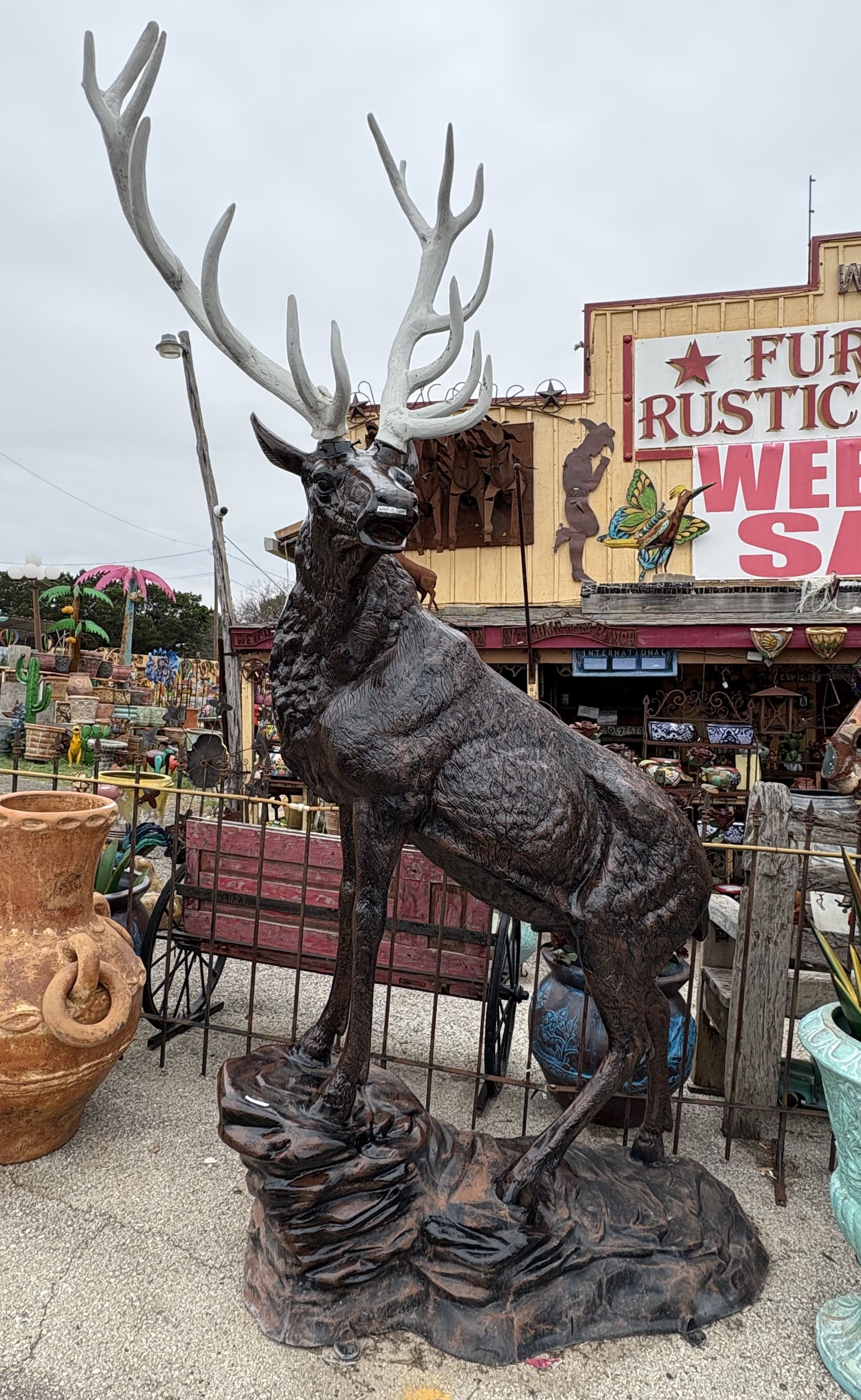 A large, dark brown elk statue with large white antlers stands outdoors in front of a rustic store.