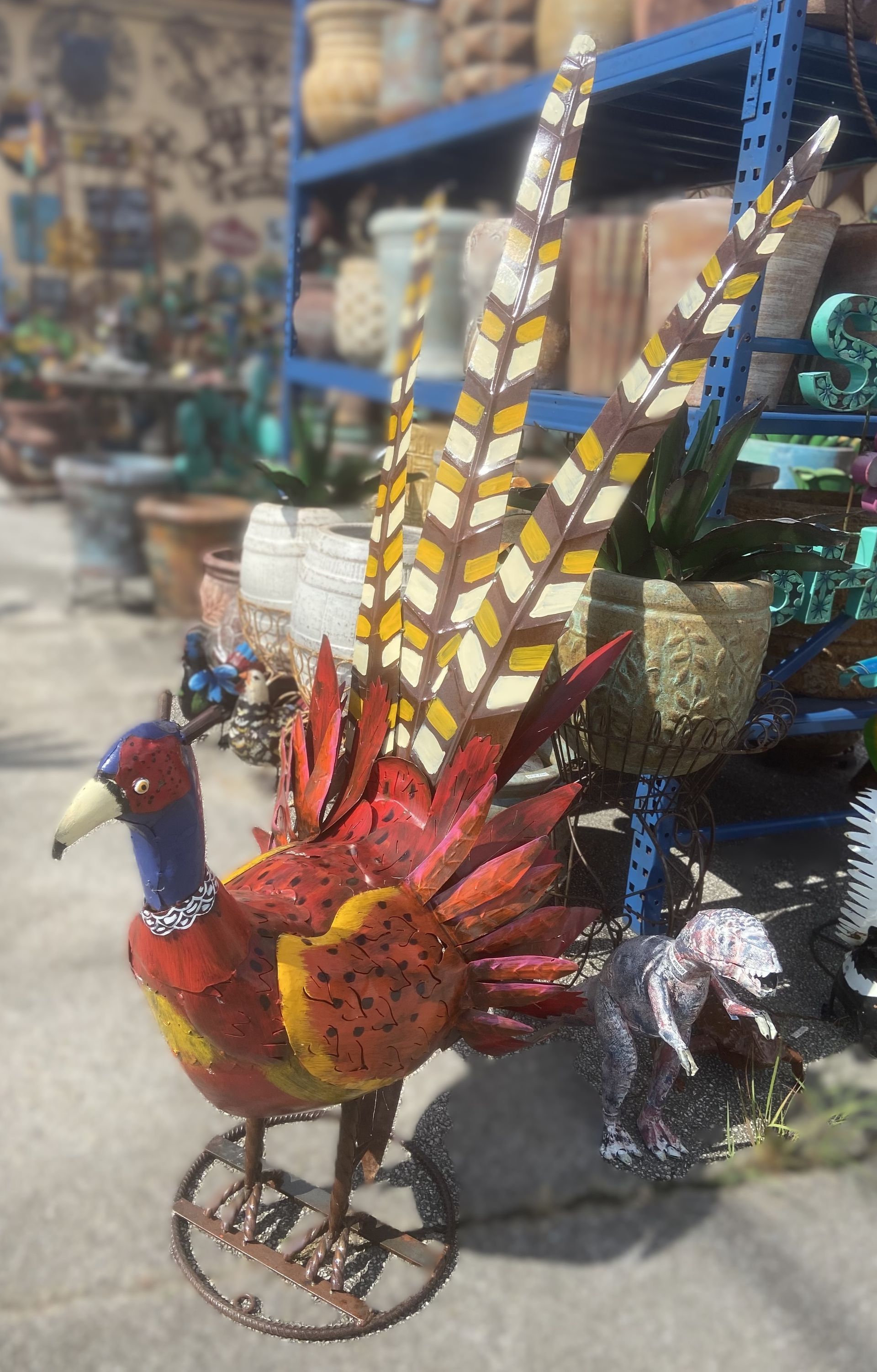 Metal pheasant garden sculpture with a vibrant red, yellow, and brown color scheme on wheels, displayed in an outdoor garden center.