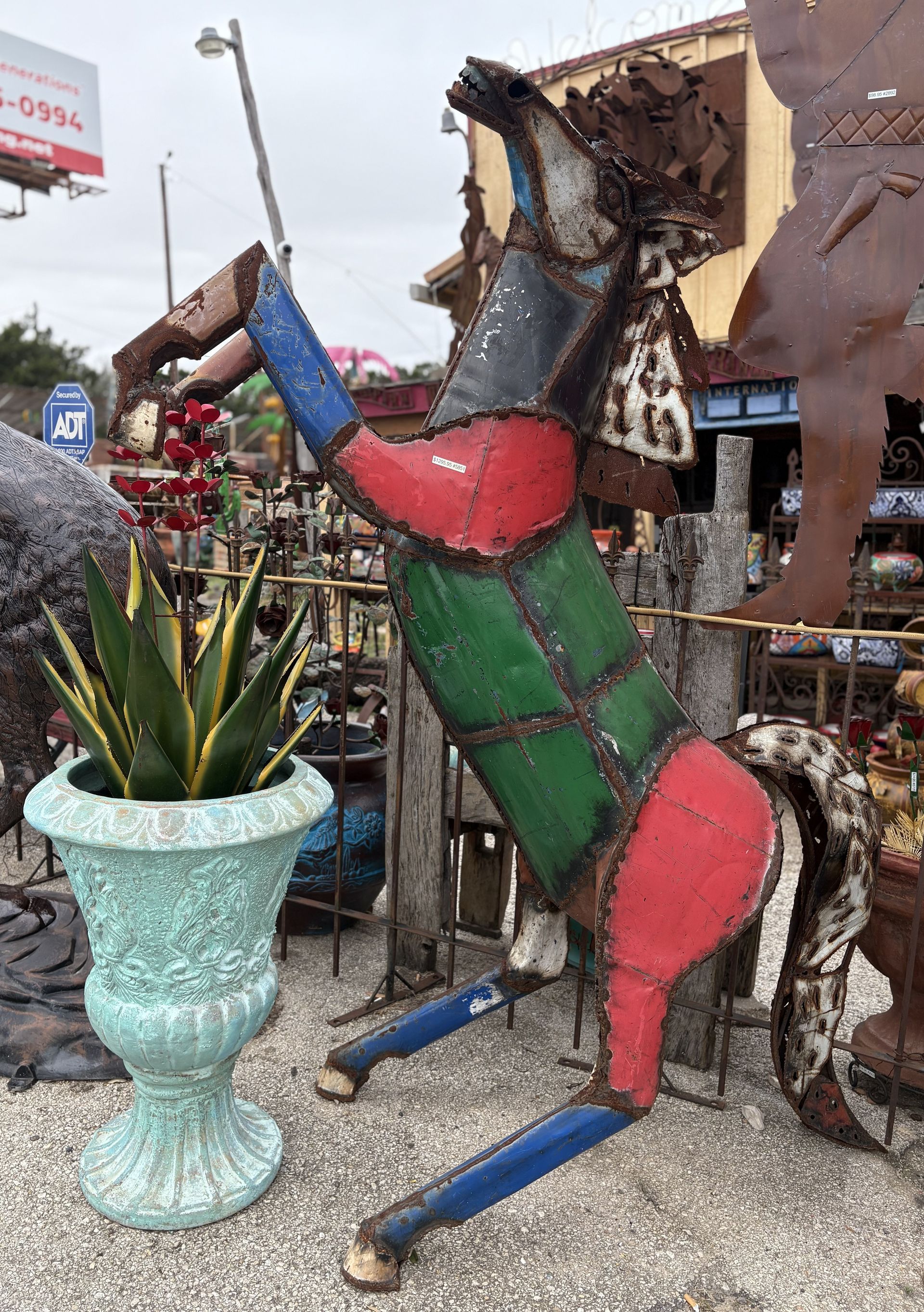 A colorful metal horse sculpture rearing up, next to a turquoise planter with a plant in an outdoor shop.