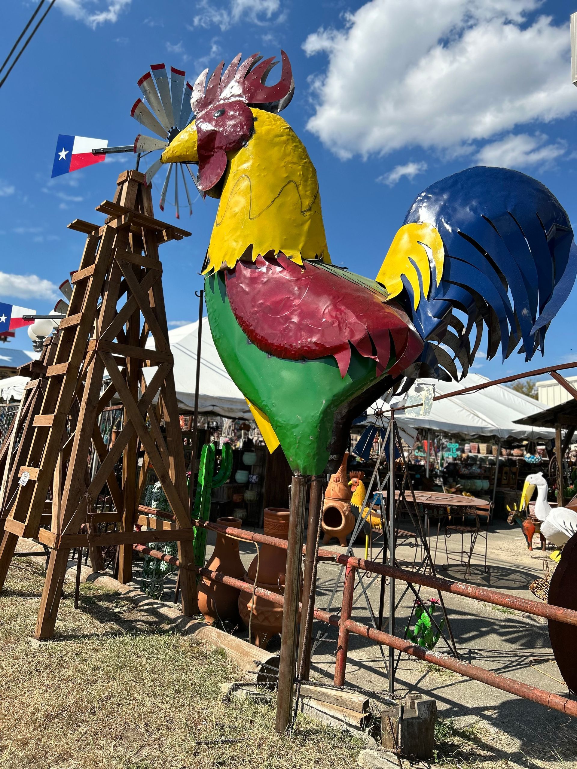 A large, colorful metal rooster statue stands outdoors with a Texas flag windmill in the background.