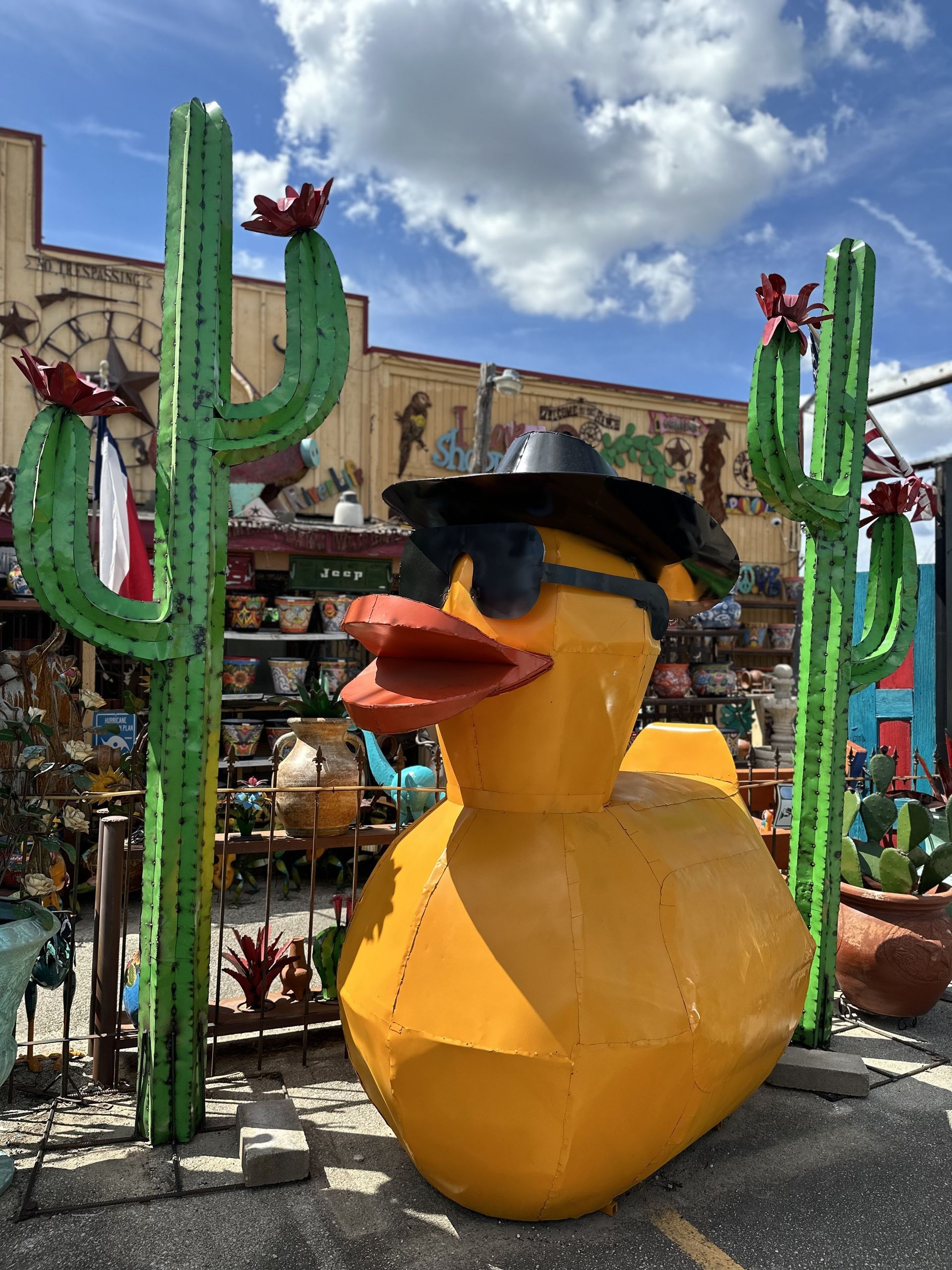 Large yellow metal duck wearing sunglasses and a hat, in front of a building with metal cactus decorations under a blue sky.