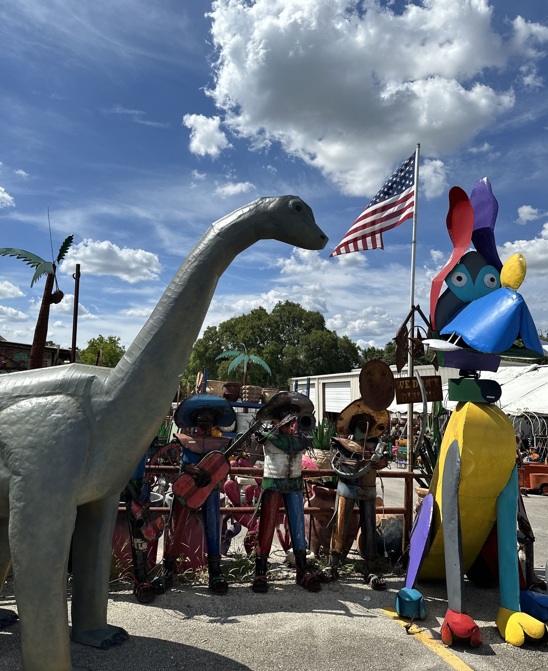 Outdoor scene with metal art sculptures: a dinosaur, a colorful bird, and musicians. An American flag waves in the background.