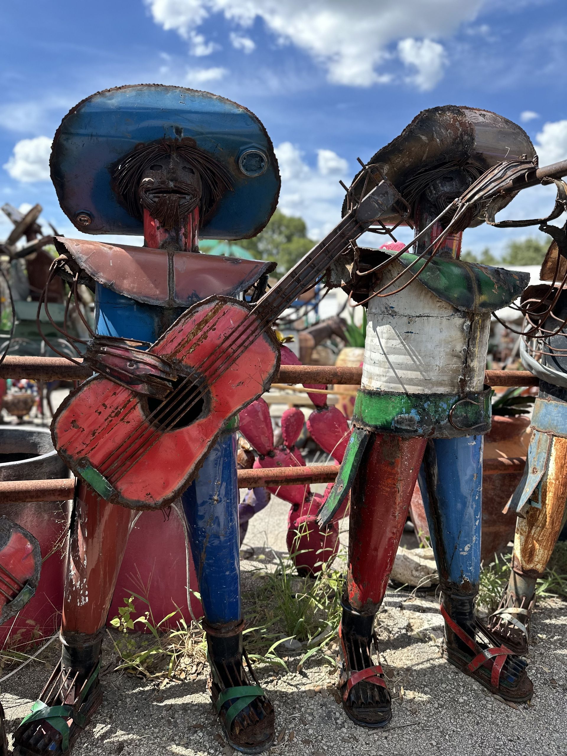 Metal sculptures of mariachi musicians playing guitar and violin, standing outdoors under a blue sky. The figures are colorful with blue, red, and green.