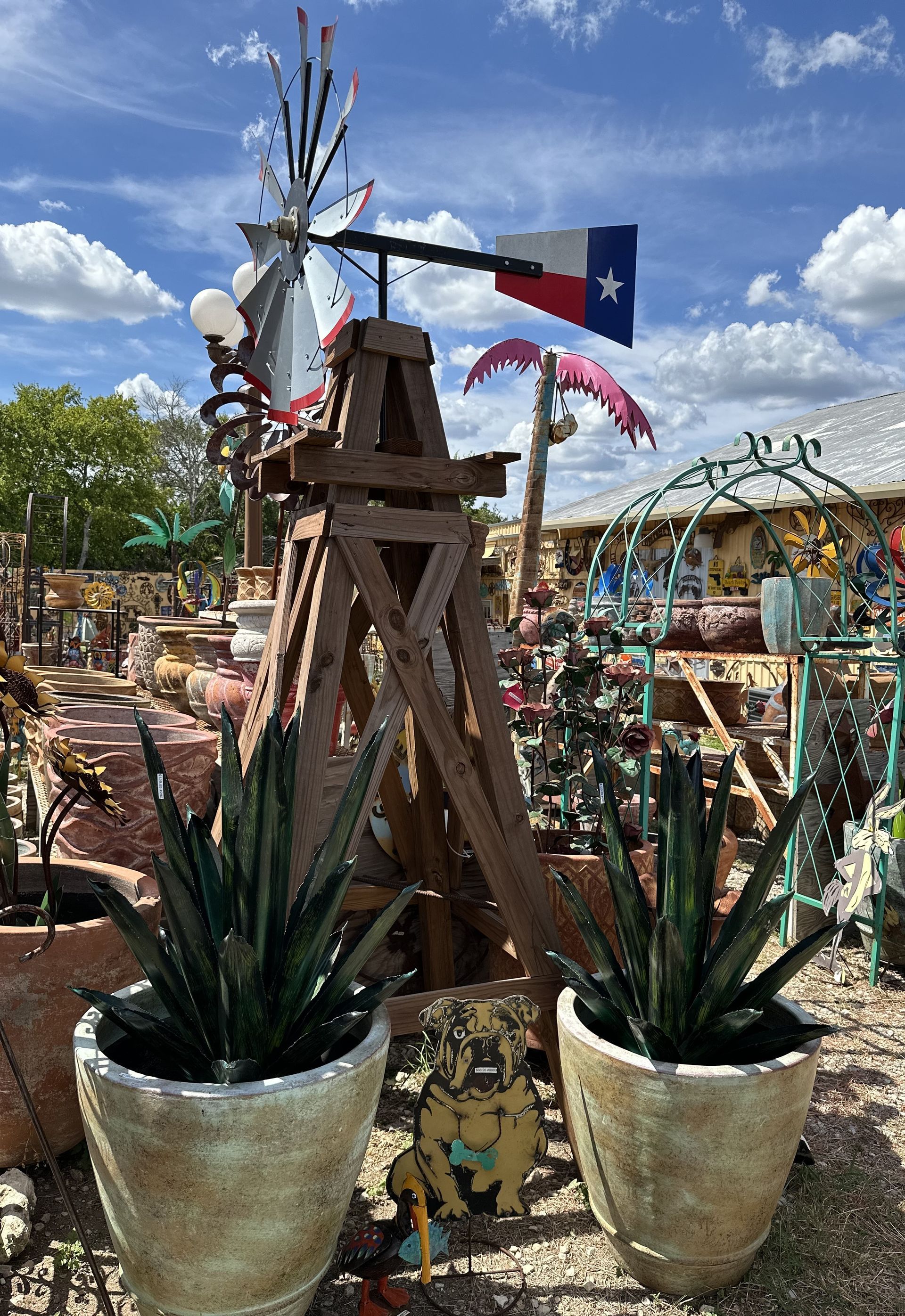 Two potted snake plants flank a wooden windmill with a Texas flag, set in a colorful garden under a blue sky.