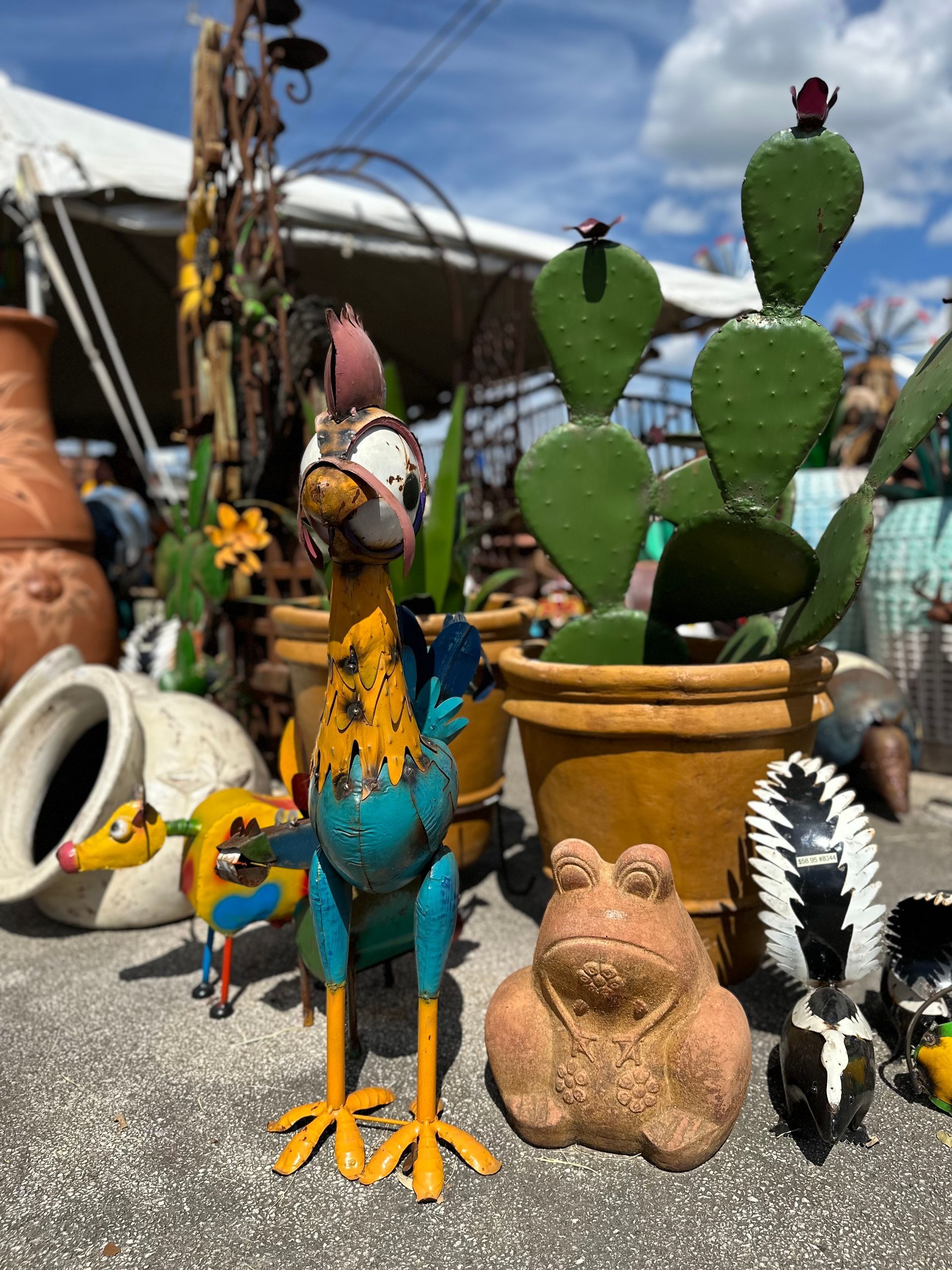 Metal rooster and cactus garden sculptures, along with a frog and skunk, in a garden center setting.