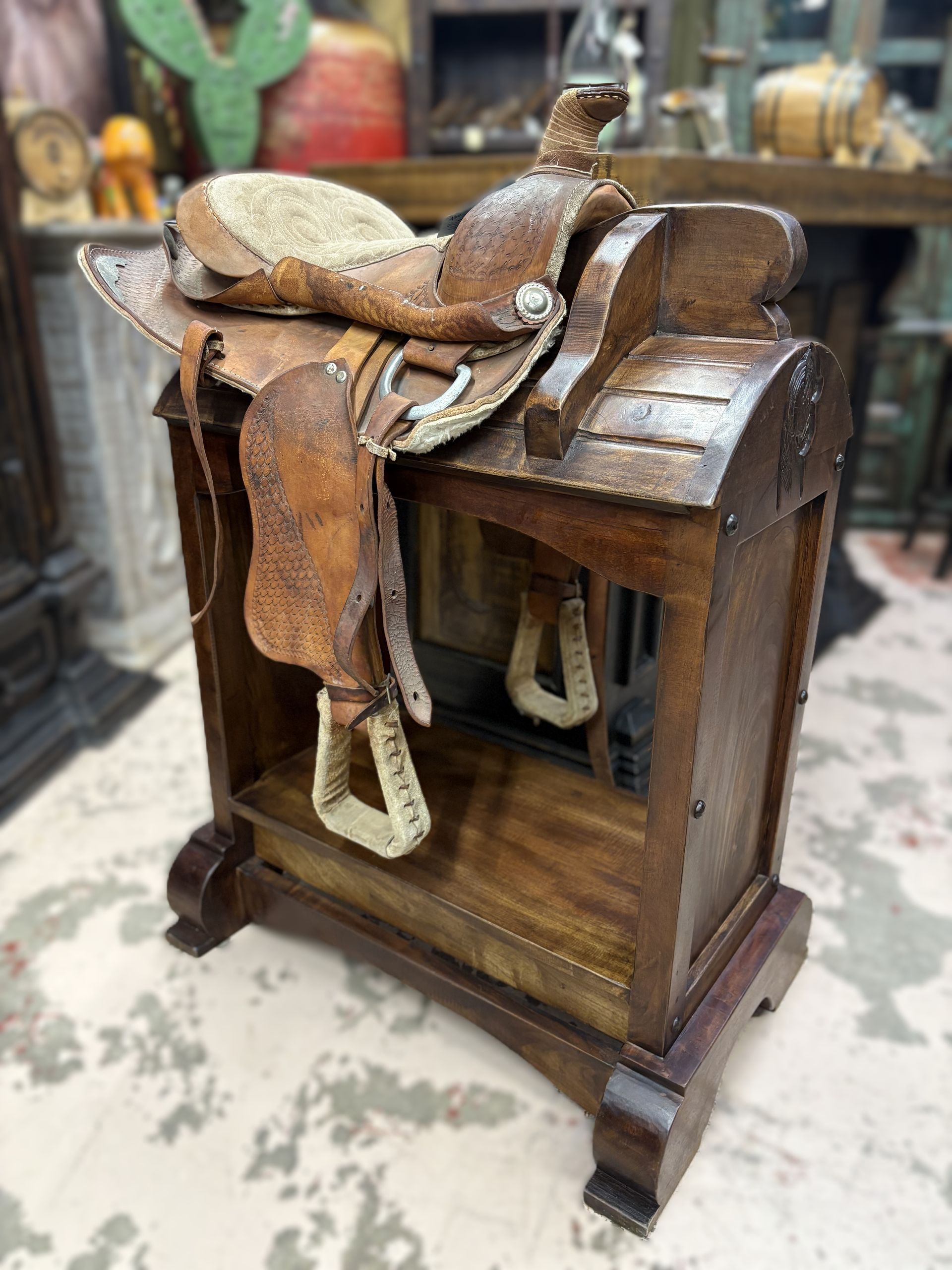 A wooden saddle stand holding a leather saddle with stirrups, set in a rustic indoor setting.