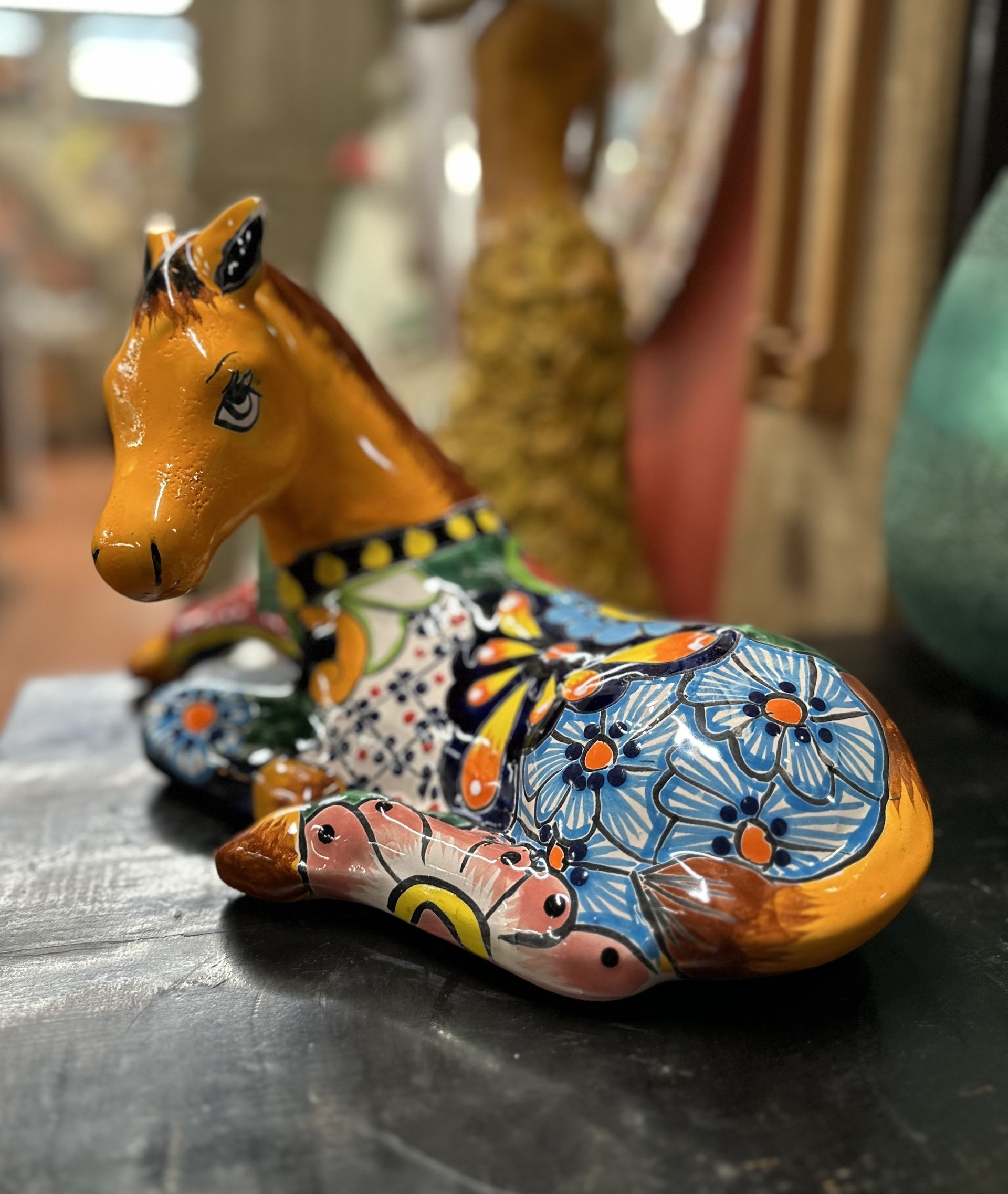 Ceramic horse figurine with colorful floral patterns, resting on a dark surface. It is displayed in an interior setting.