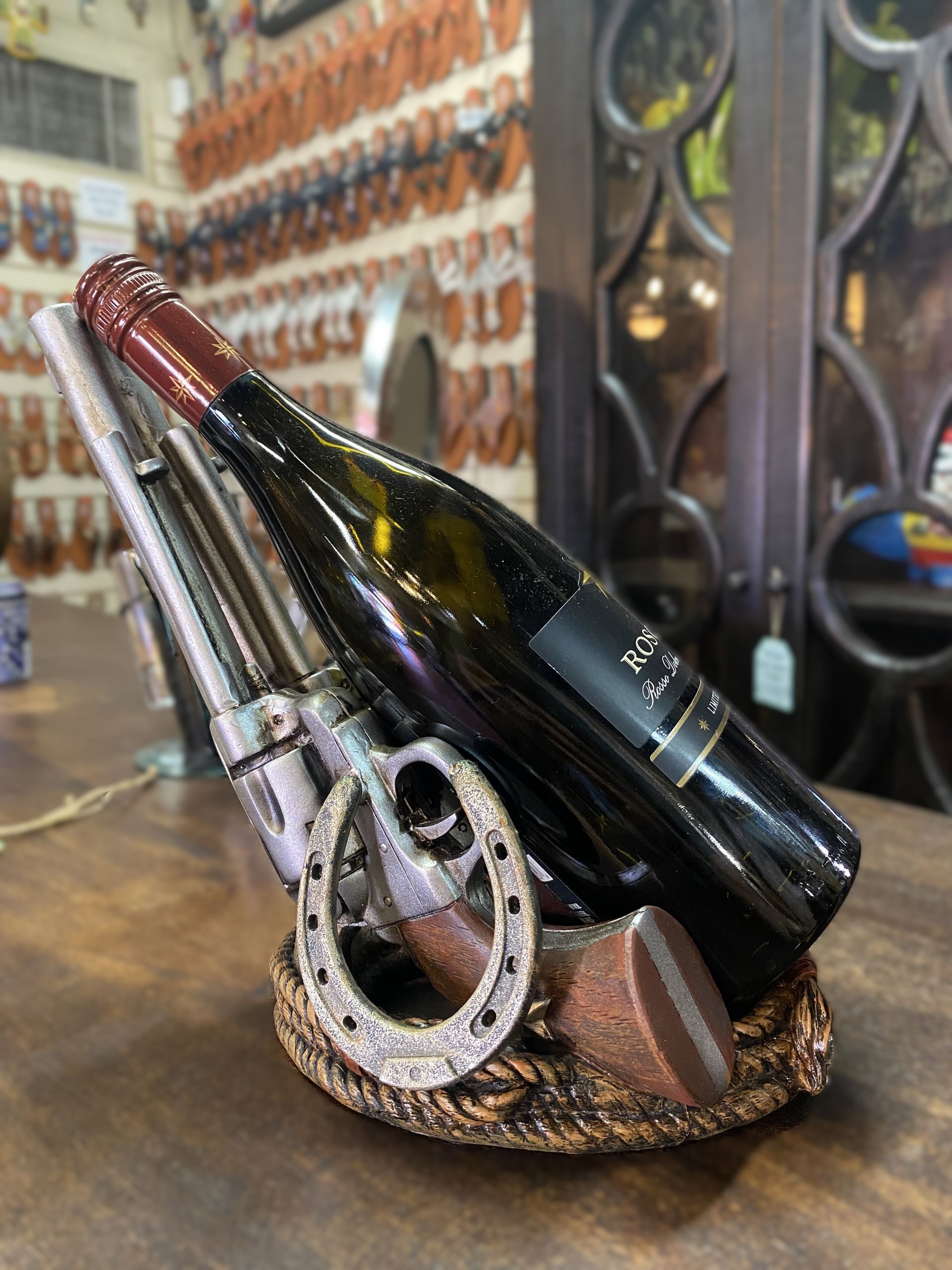 A bottle of red wine in a Western-themed holder featuring a pistol, horseshoe, and rope detail, on a wooden surface.