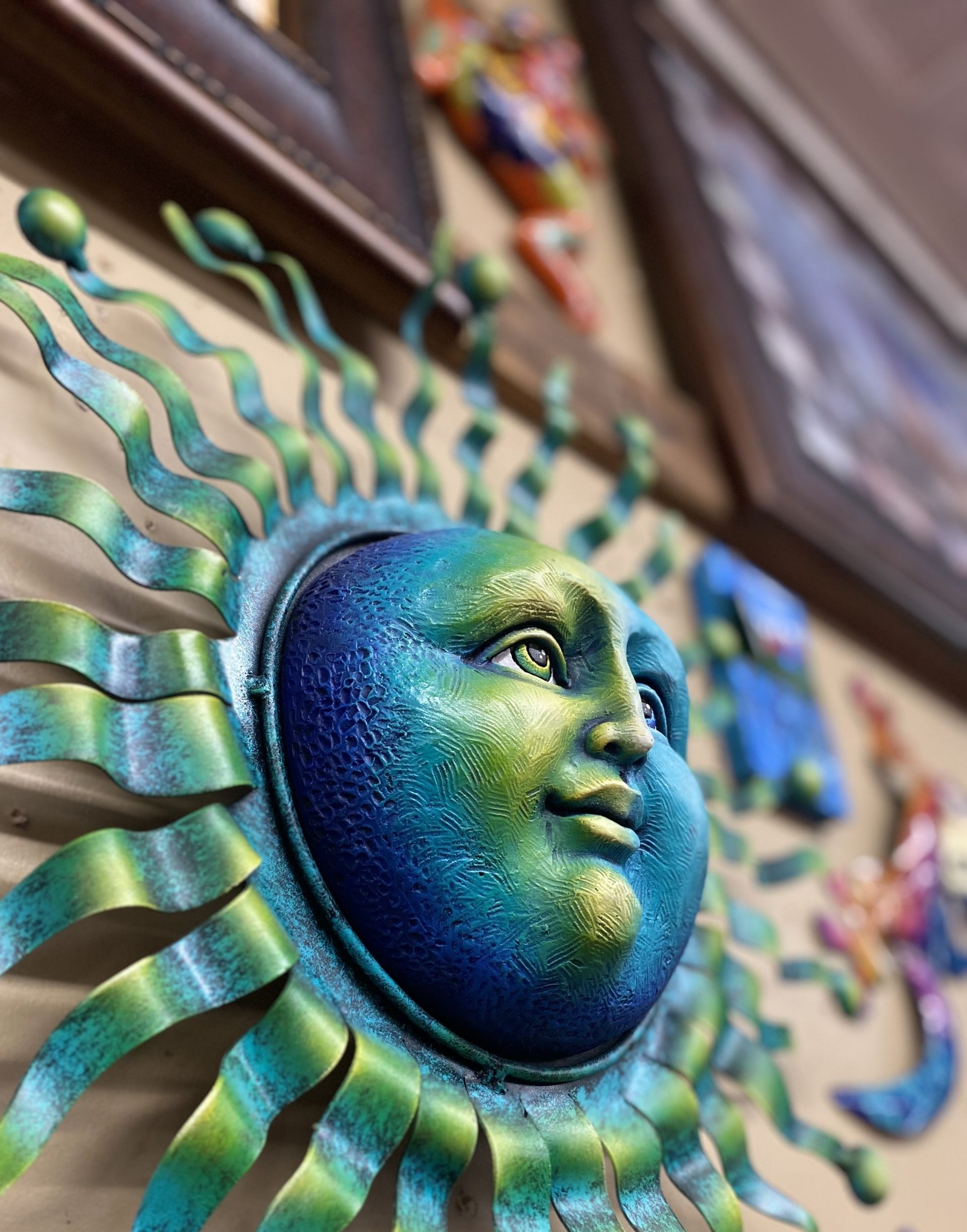 Metal sun wall decoration with blue and green hues, set against a tan wall with framed artwork.