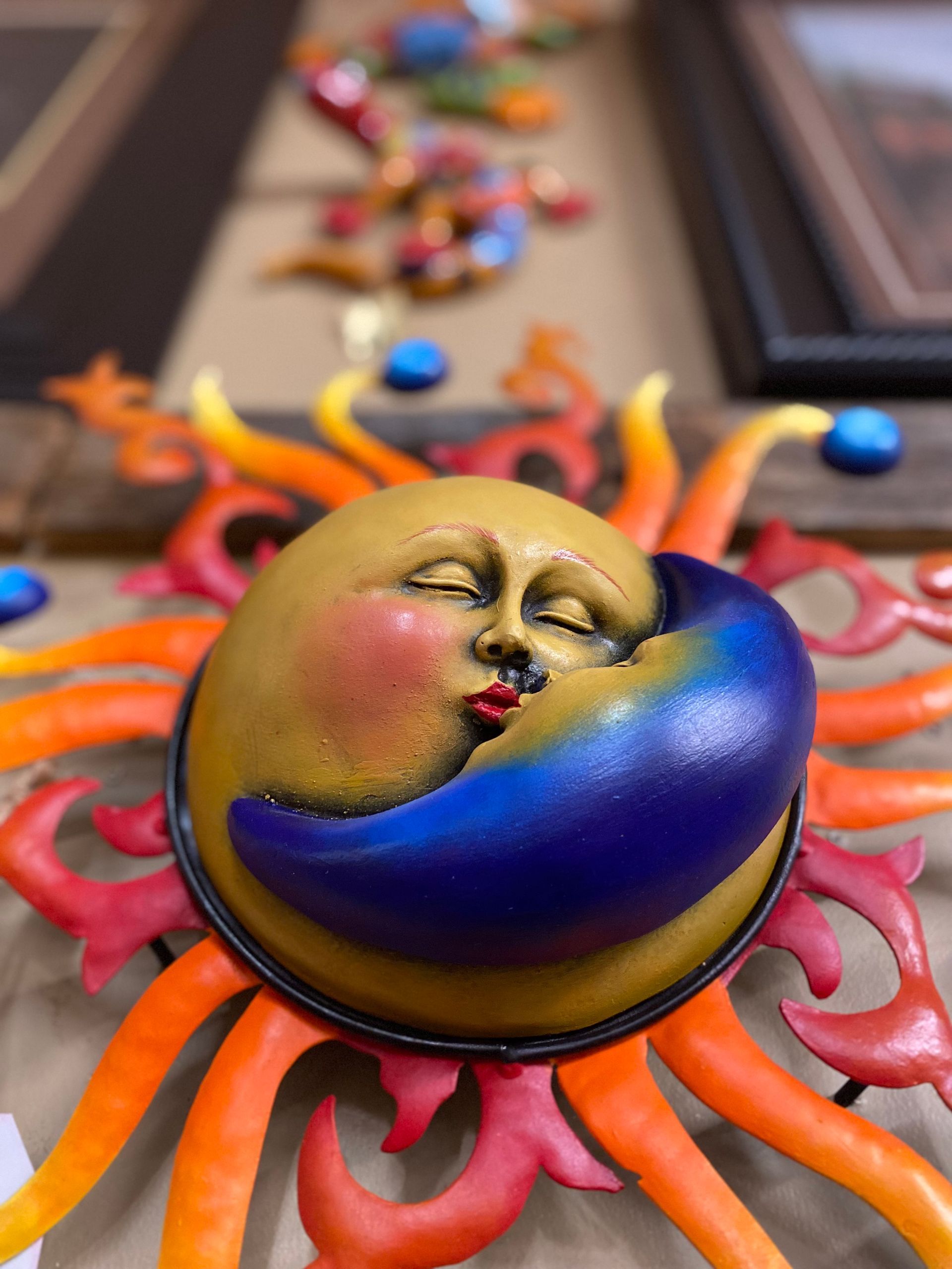 A whimsical sun face with closed eyes and a kissing crescent moon, surrounded by colorful rays; mounted on a wall.