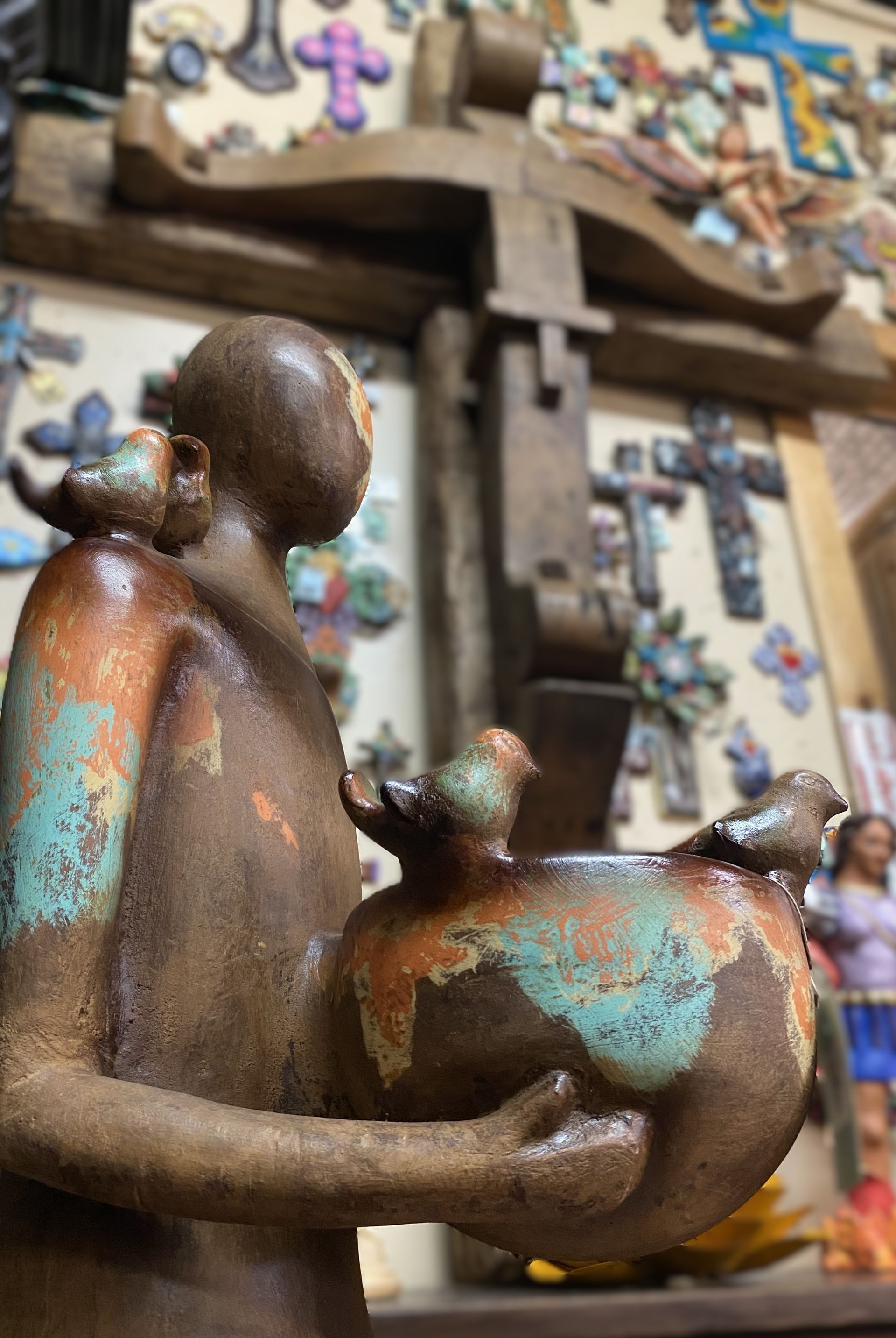 Brown ceramic figure holding a bowl with birds; figure has a bird on its shoulder. Background shows decorative crosses.