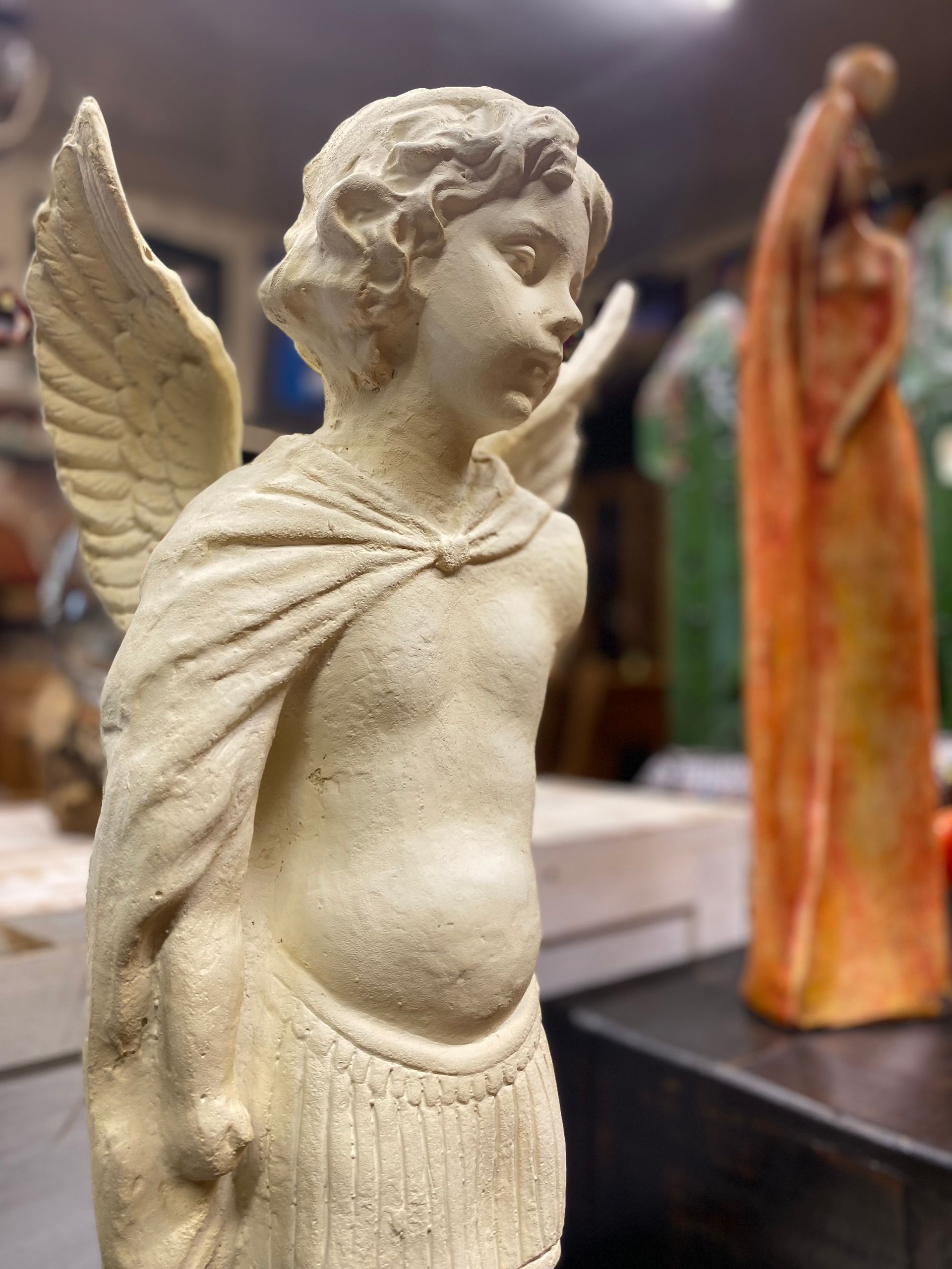 A stone cherub statue with wings and a draped shawl, standing near a tall, slender, orange figure.