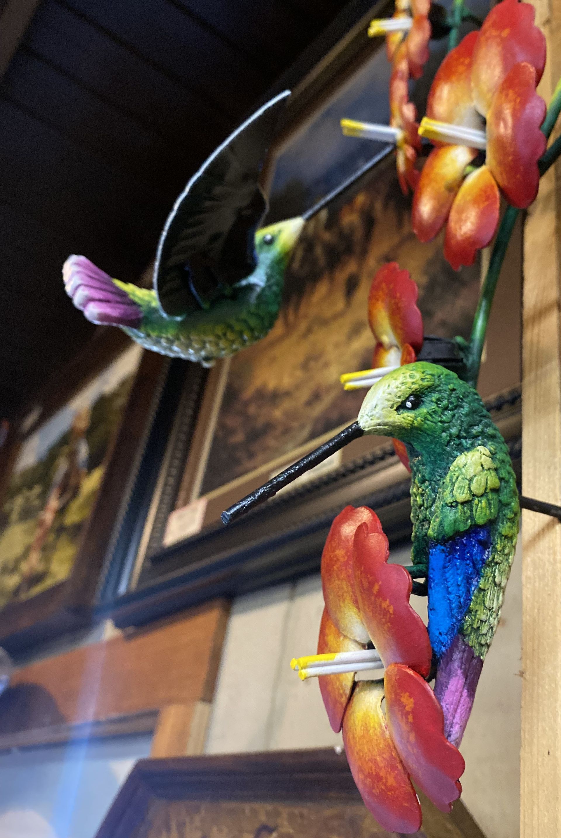 Two colorful hummingbird statues feeding on red and yellow flowers. One bird is in flight; both have long beaks.