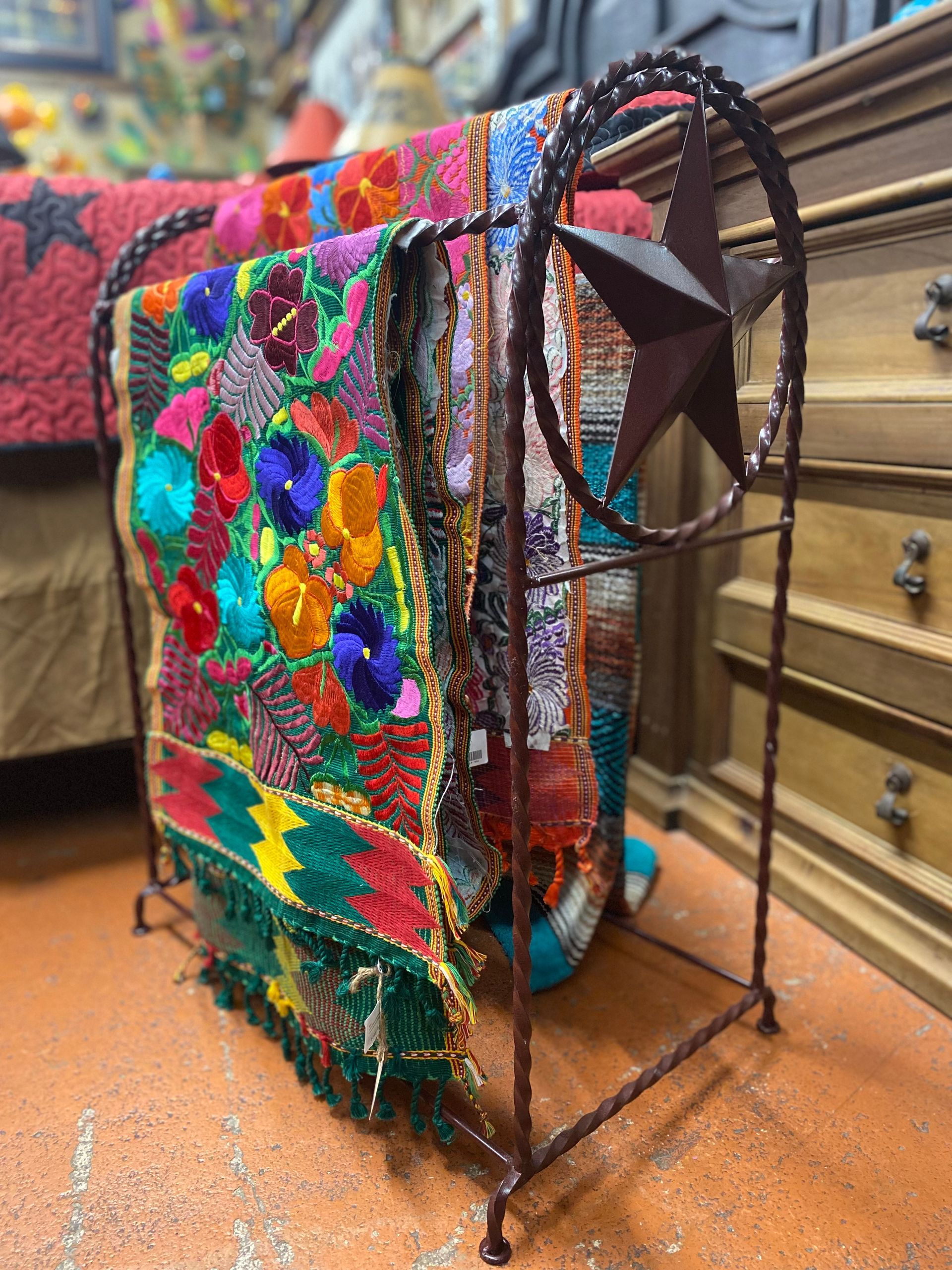 A rustic metal blanket rack displaying colorful embroidered textiles. The rack features a star design on the side, near a wooden dresser.