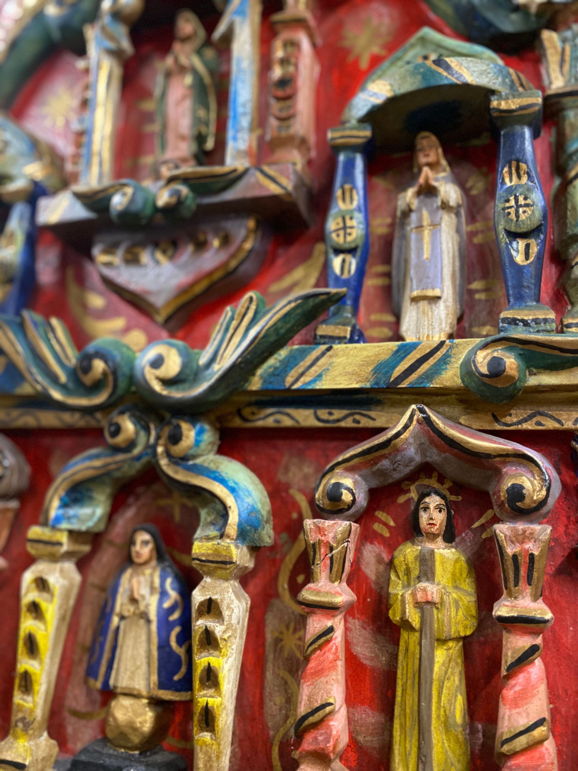 Close-up of a brightly painted, ornate wooden religious carving. Figures of saints are framed by colorful architectural details.