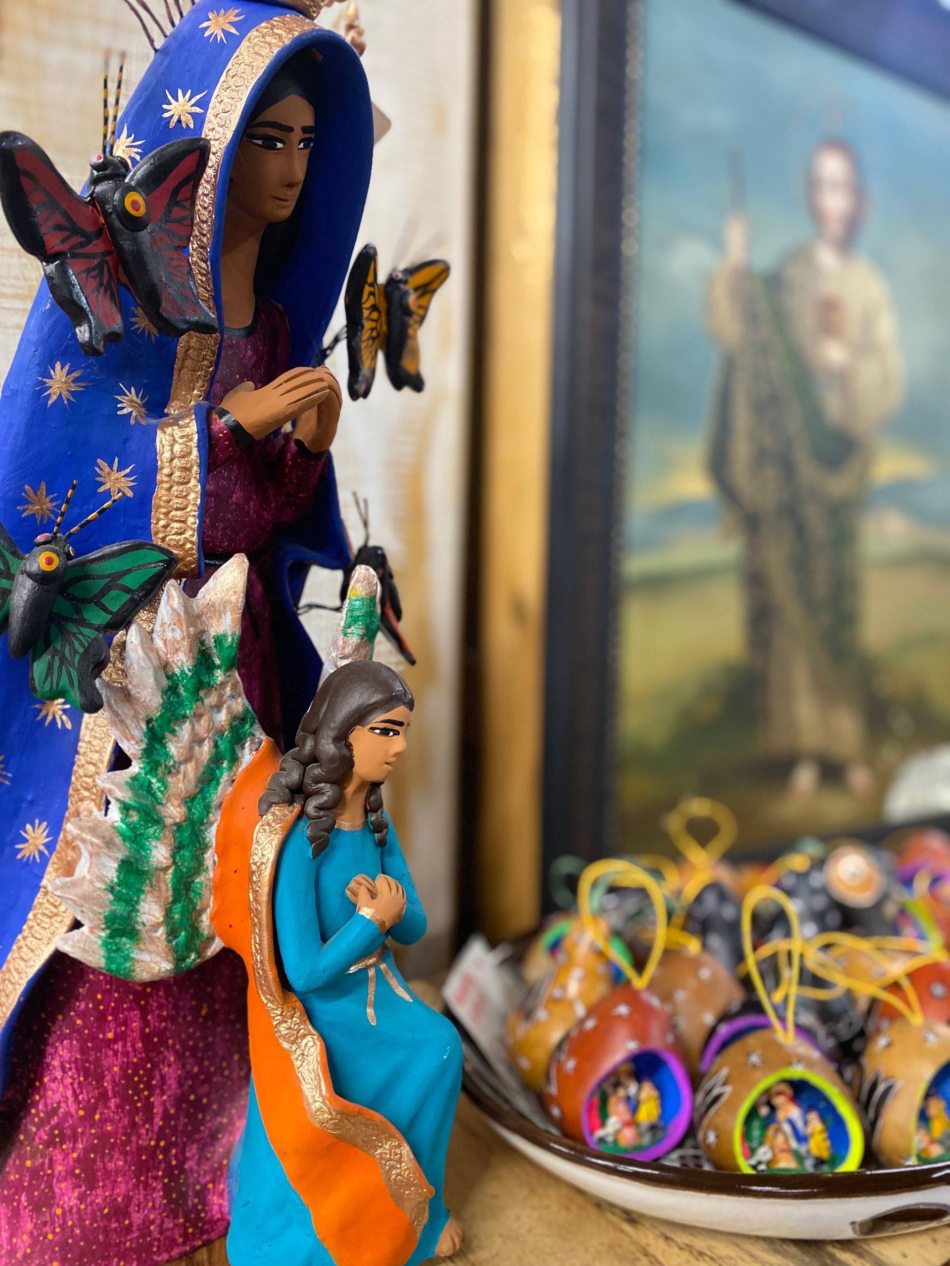 A display featuring colorful religious figures and decorations, including statues of saints, with a blurred painting in the background.