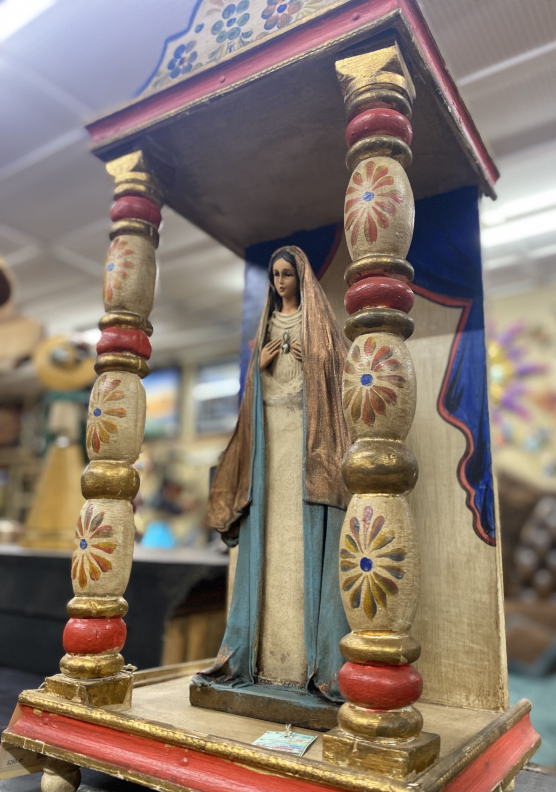 Statue of the Virgin Mary inside a decorative, painted wooden shrine. The statue is wearing a blue robe with her hands clasped in front of her.
