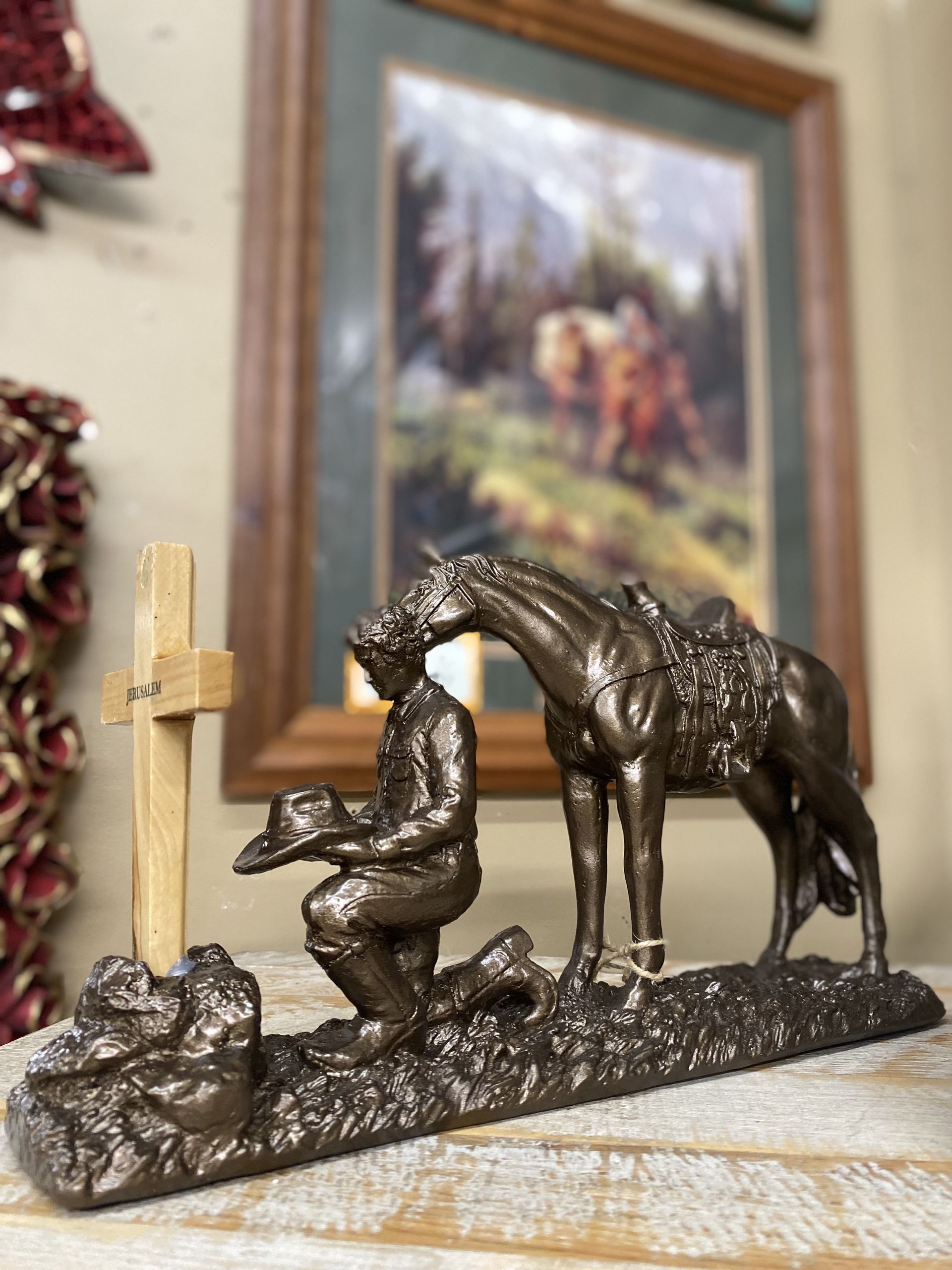 Bronze statue of a kneeling cowboy praying by a horse, with a wooden cross nearby. Behind them is a framed painting of horses in a landscape.