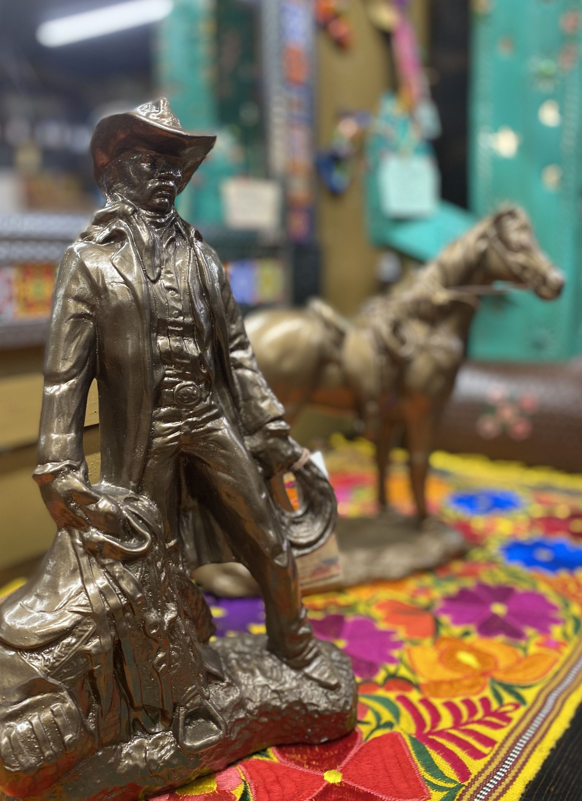Bronze cowboy statue holding a saddle, with a horse statue blurred in the background, on a colorful cloth.