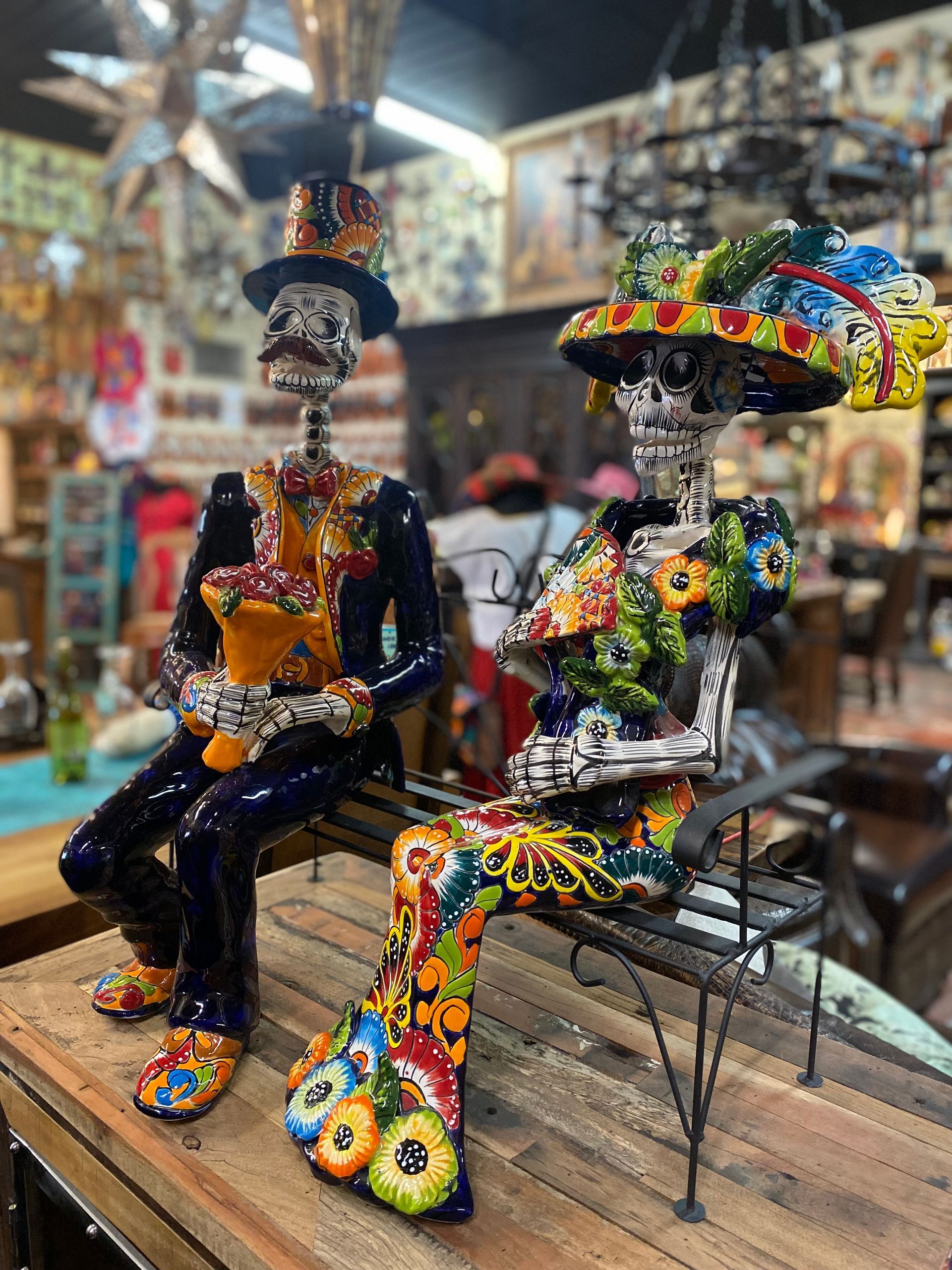 Two colorful Day of the Dead skeleton figures, a man in a top hat holding a flower and a woman with a floral hat, seated.