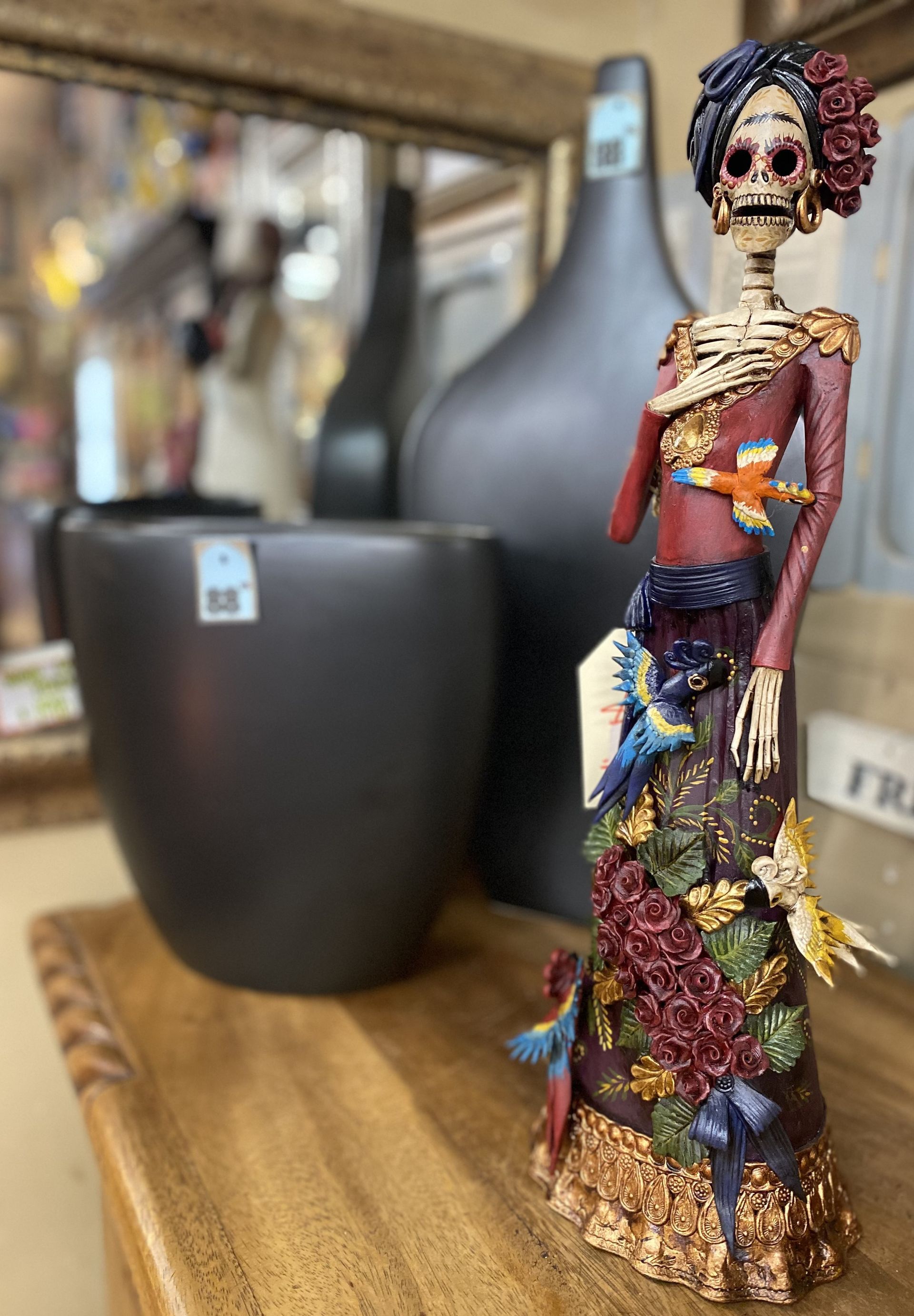 A Day of the Dead skeleton figurine in a colorful dress, standing on a wooden surface, with black vases in the background.