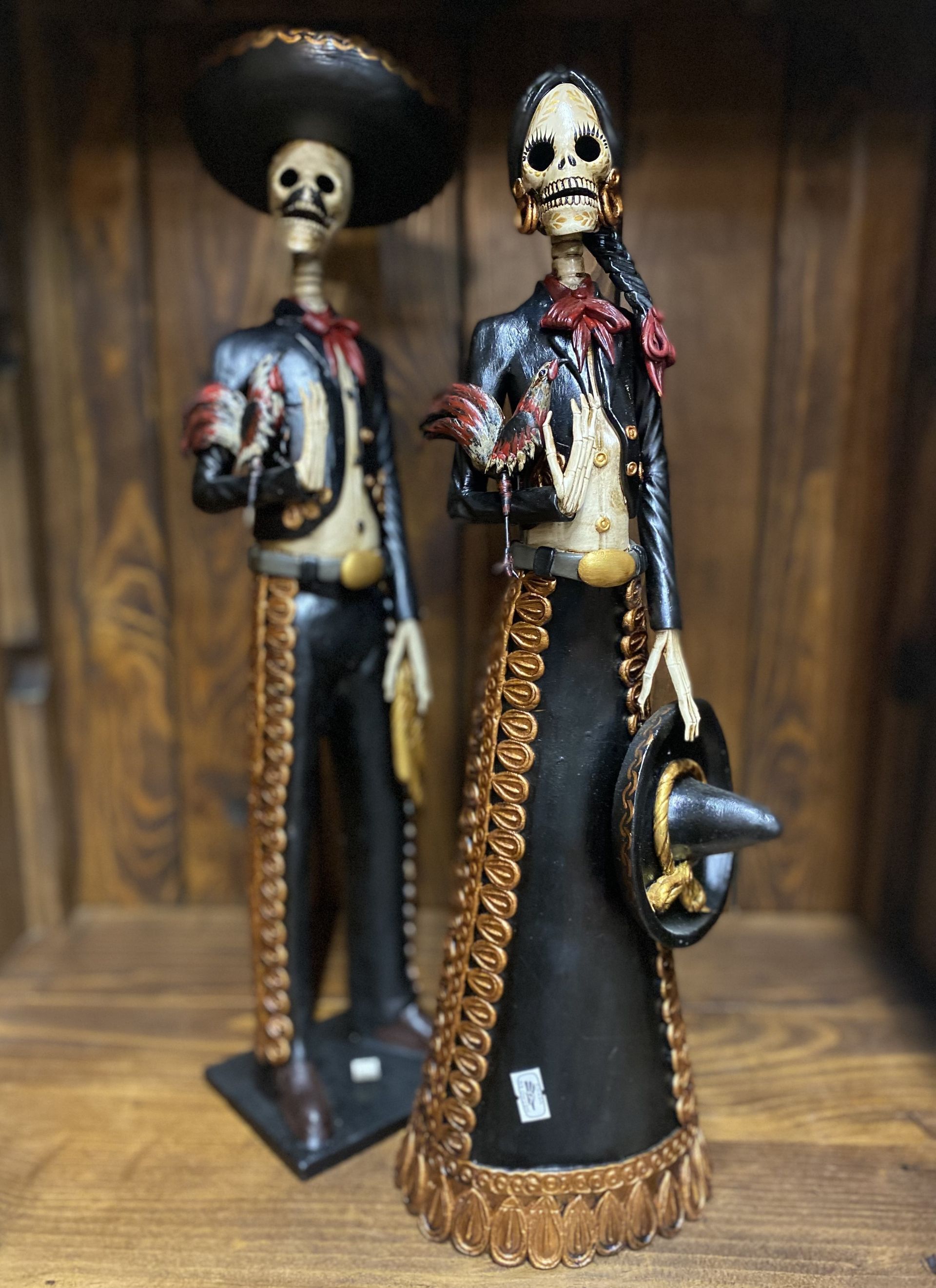 Two skeletal figures, a man and woman in traditional Mexican attire, stand before wooden planks. The man is wearing a wide-brimmed hat, while the woman holds one.