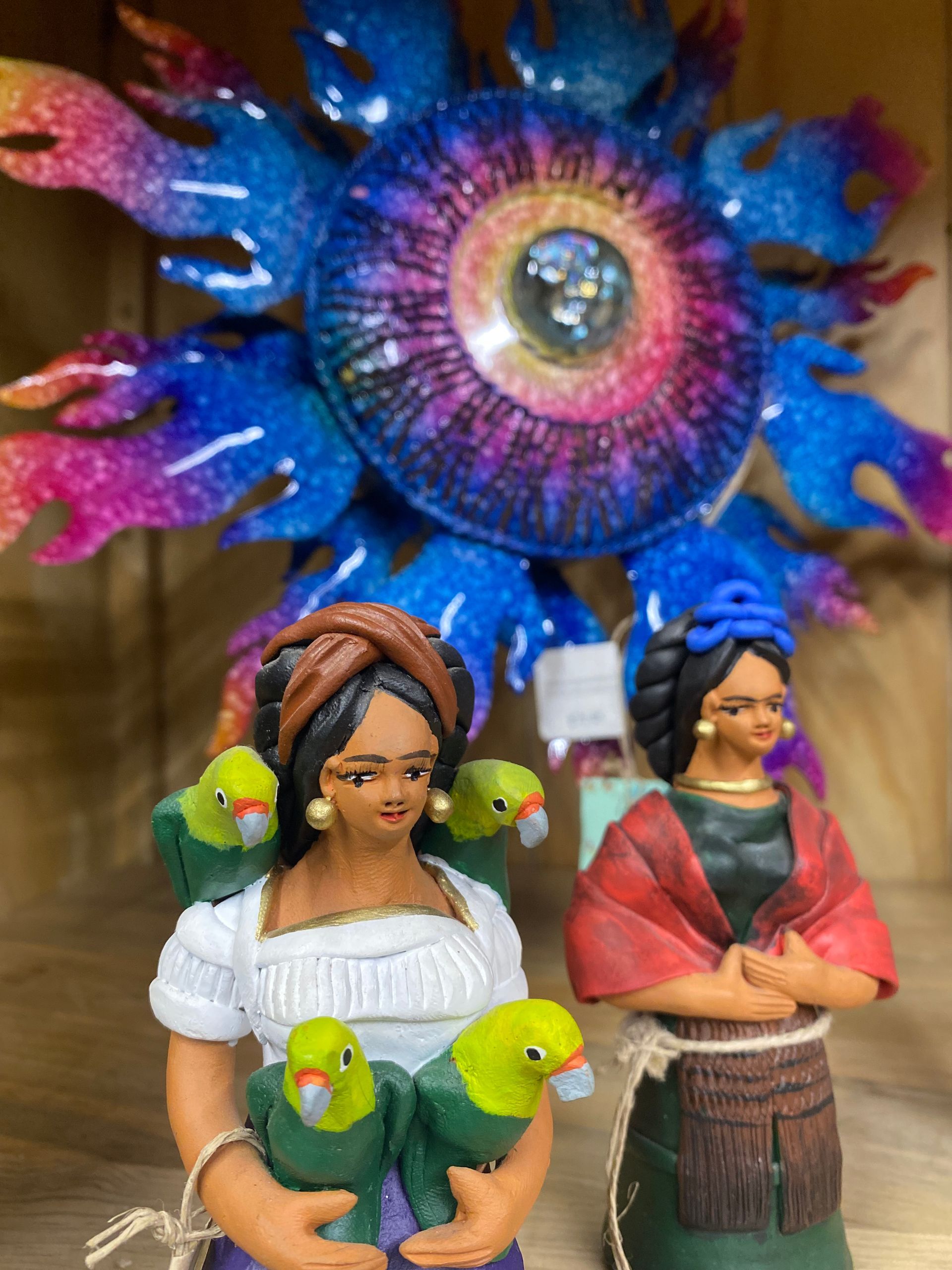 Two figurines of women with parrots in front of a colorful, sparkly sunburst. One woman has birds on her shoulders.