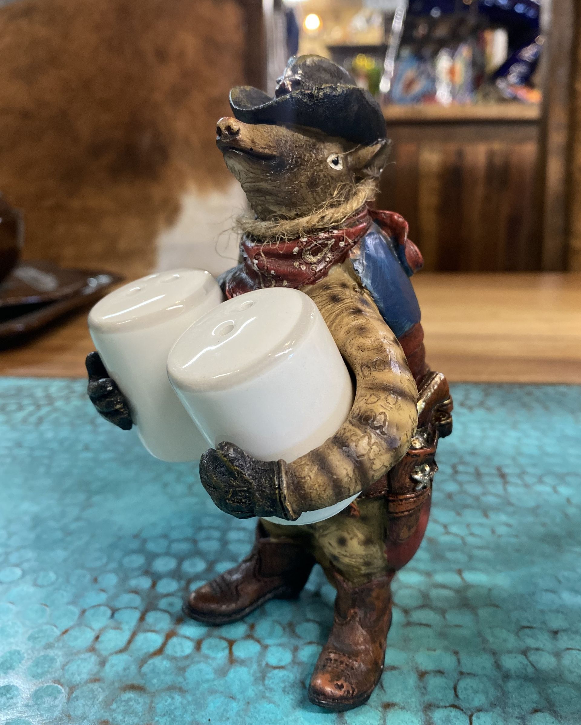 A cowboy possum figurine holding two white salt shakers on a blue textured surface. The possum wears a hat, vest, and boots.