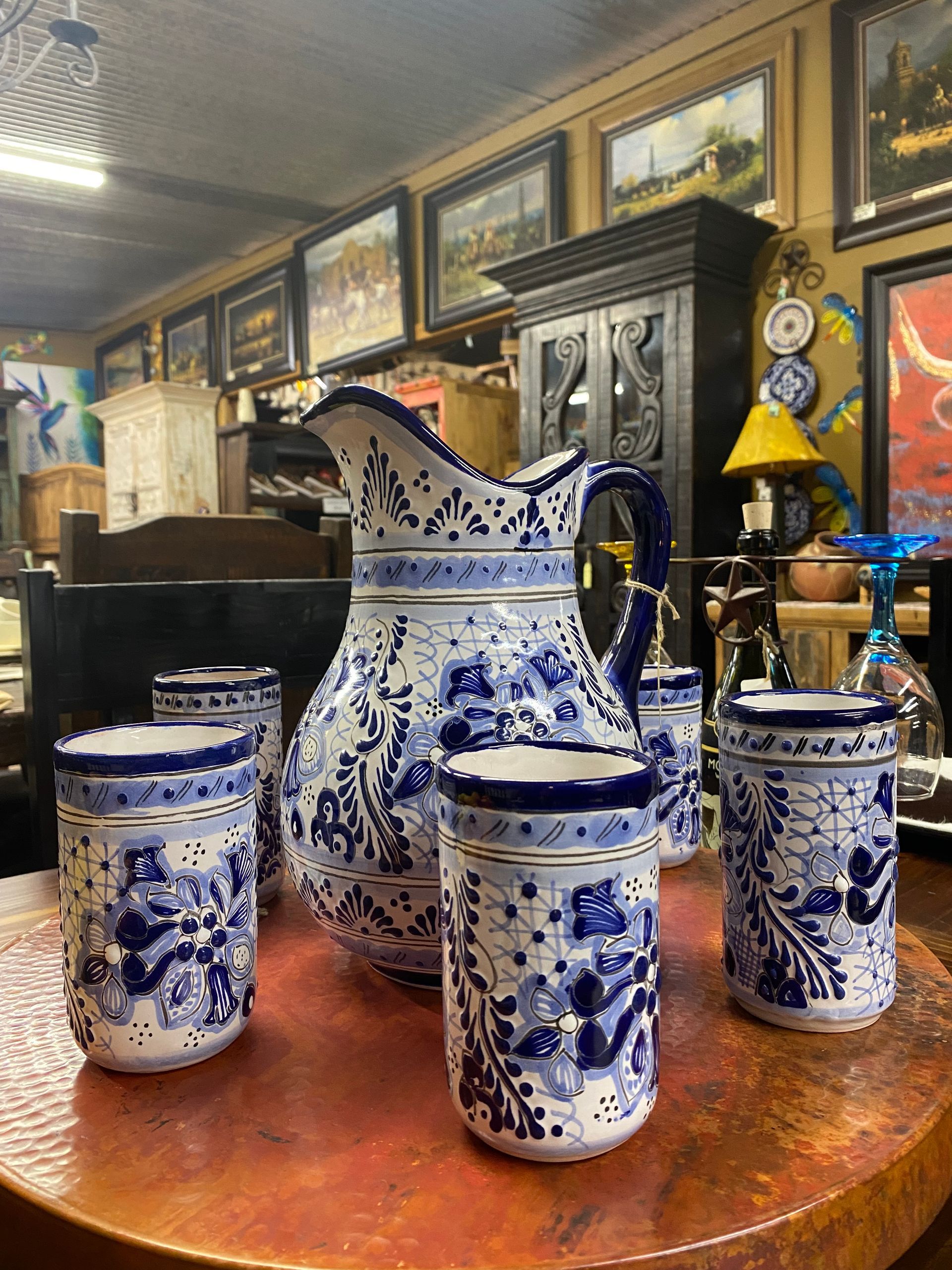 Blue and white ceramic pitcher and glasses on a copper surface, set against a backdrop of antiques and artwork in a shop.