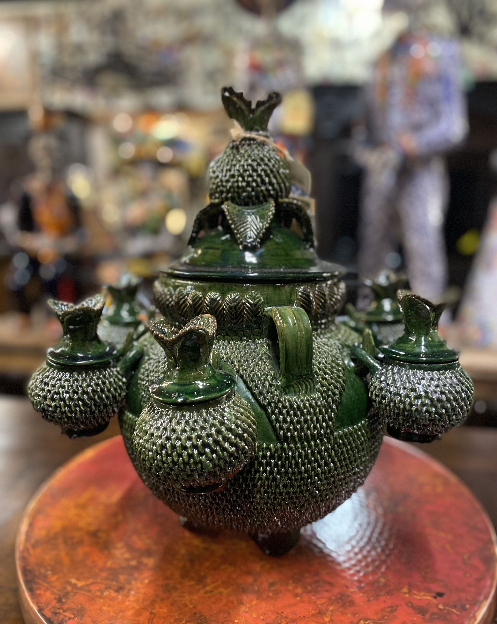 Green ceramic sculpture on a reddish-brown surface. It is round with many smaller attached vessels and ornate textured details.