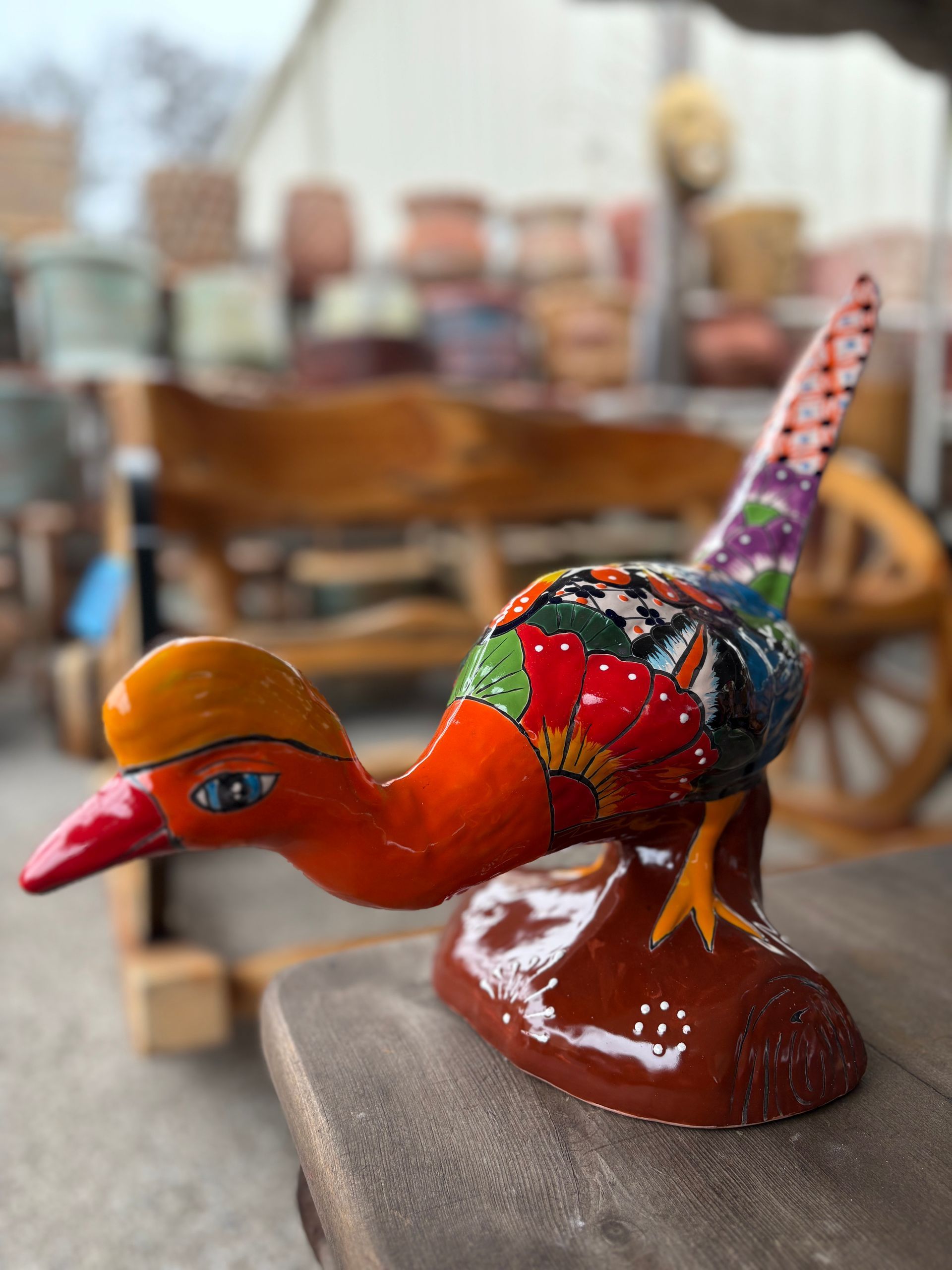 Brightly painted bird sculpture on a table outdoors. Orange head, red beak, multi-colored body, perched on a brown base.