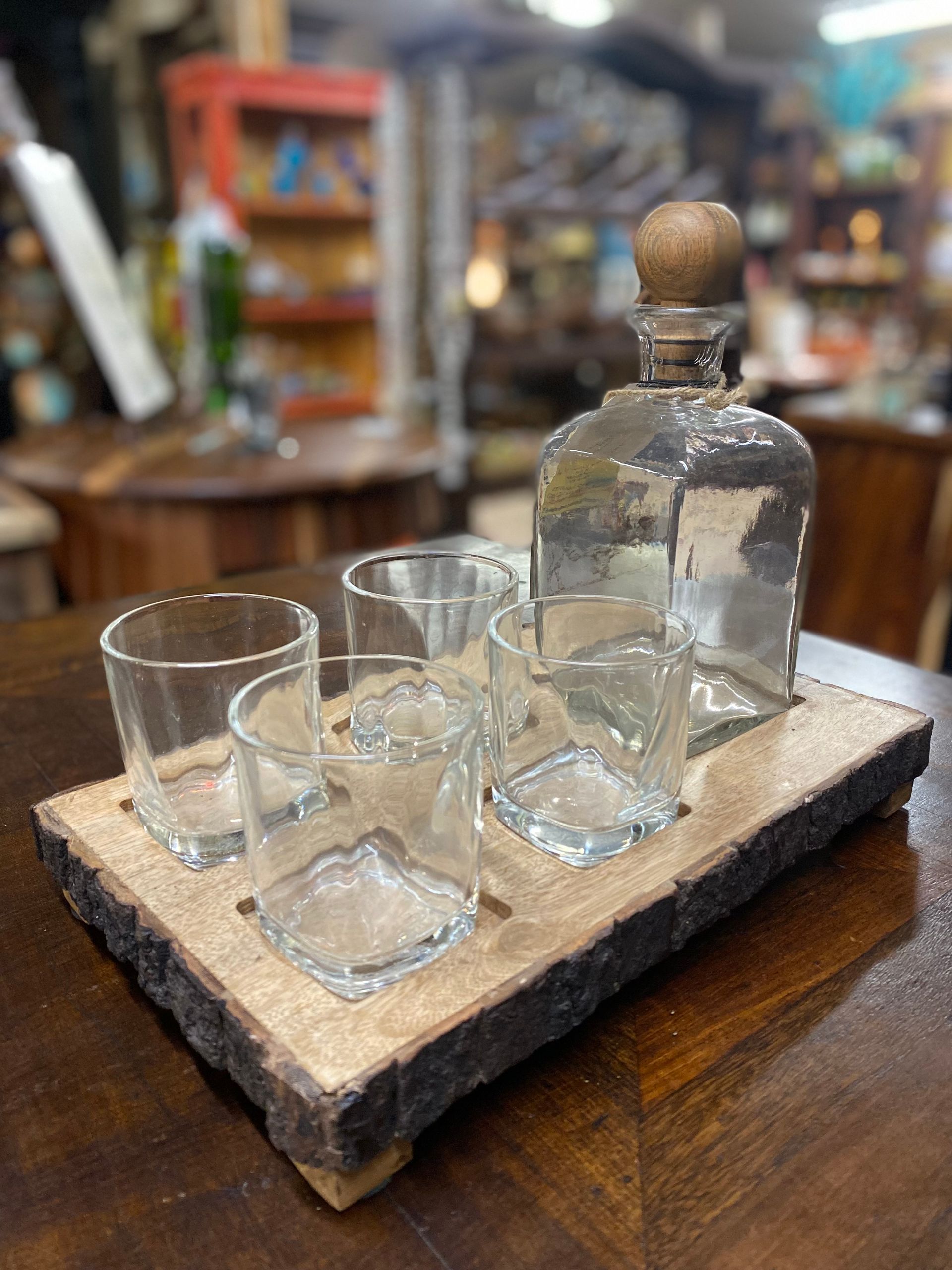 A wooden serving tray holding a tequila bottle with a cork and five empty shot glasses. The setting is indoors.