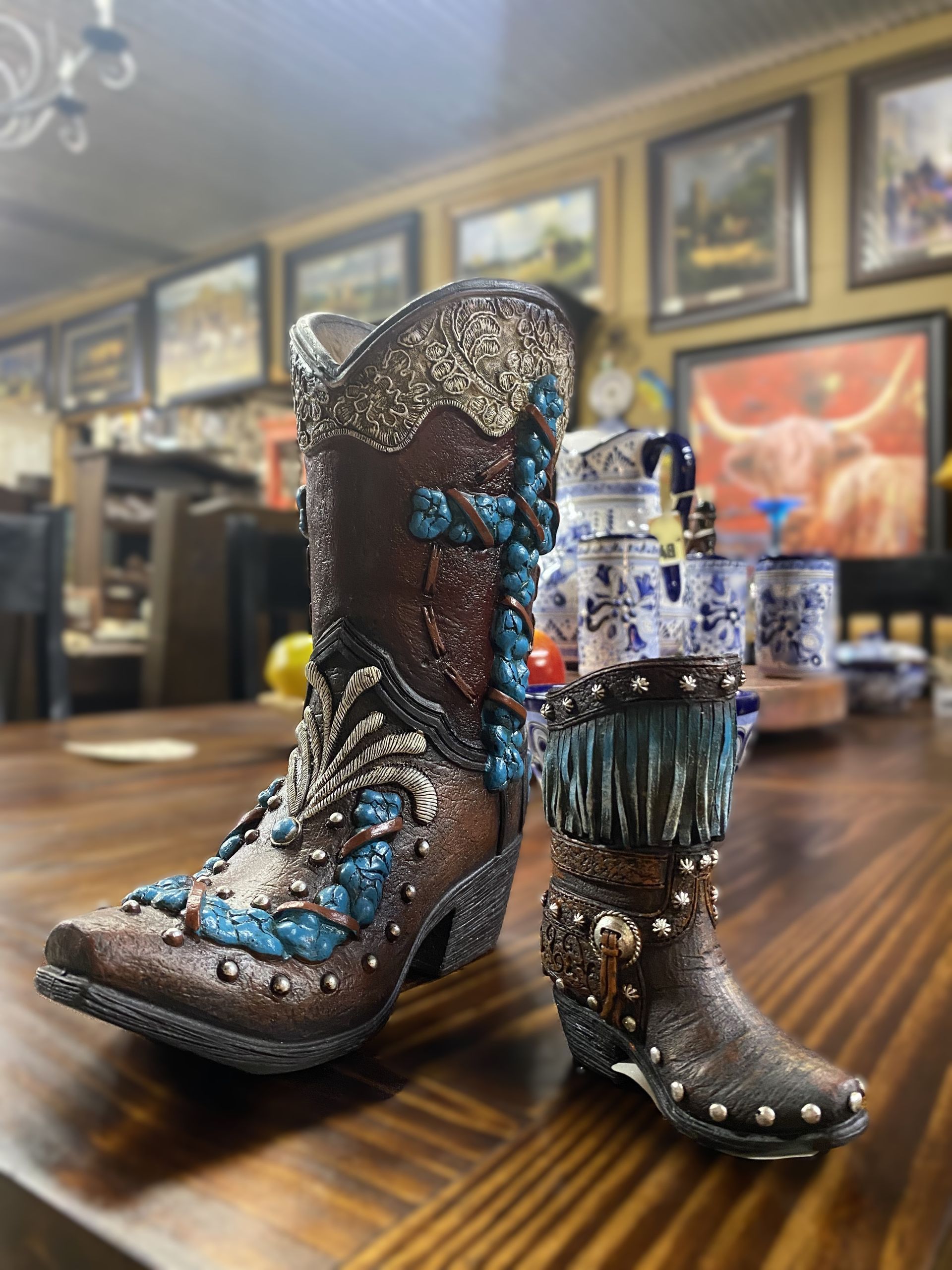 Brown and turquoise cowboy boot figurines on a wooden table. The larger boot is highly decorated with glitter and embellishments.