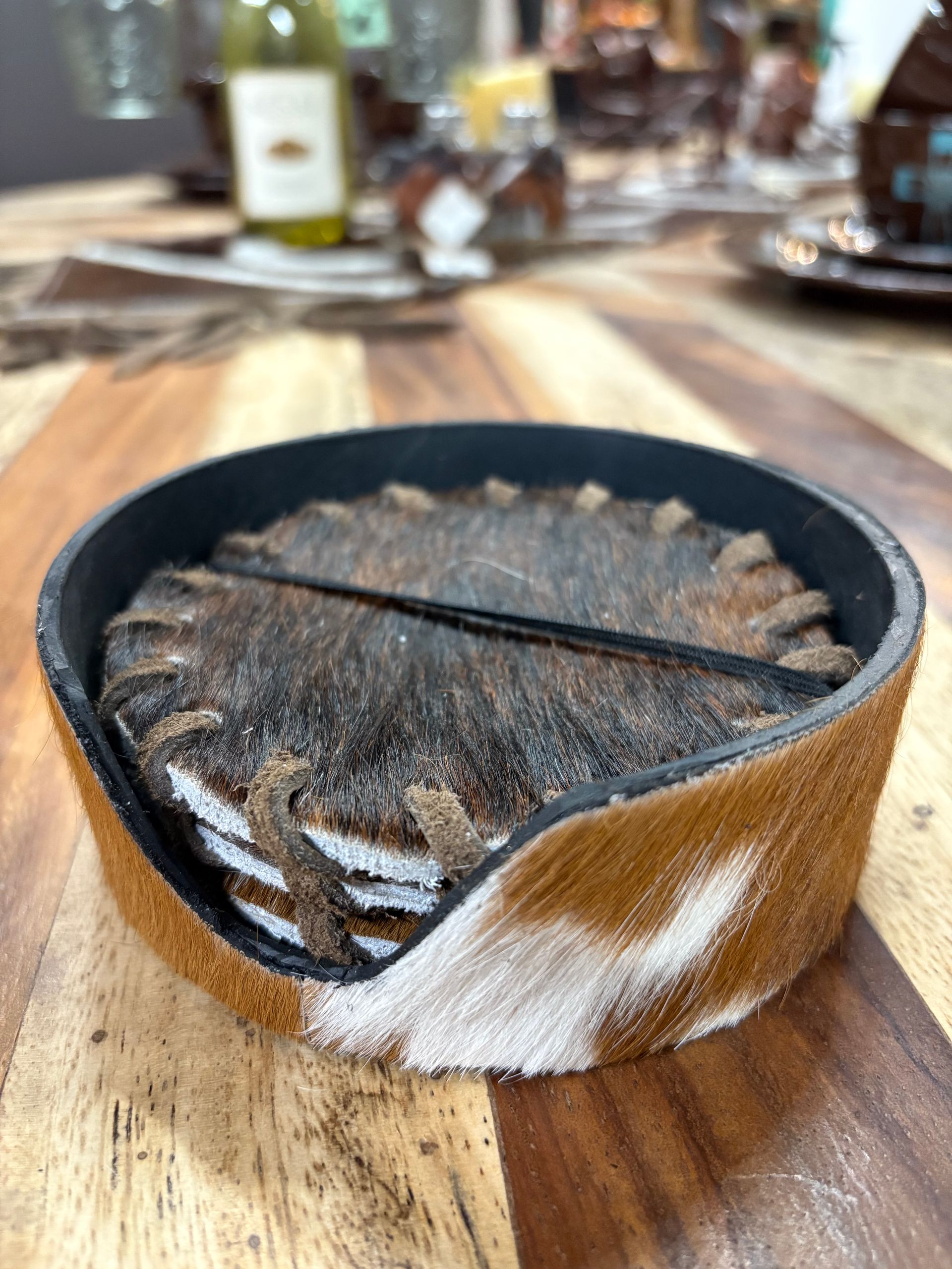 A stack of cowhide coasters in a round, leather-rimmed holder on a wood table. The coasters are brown and white with stitched detailing.