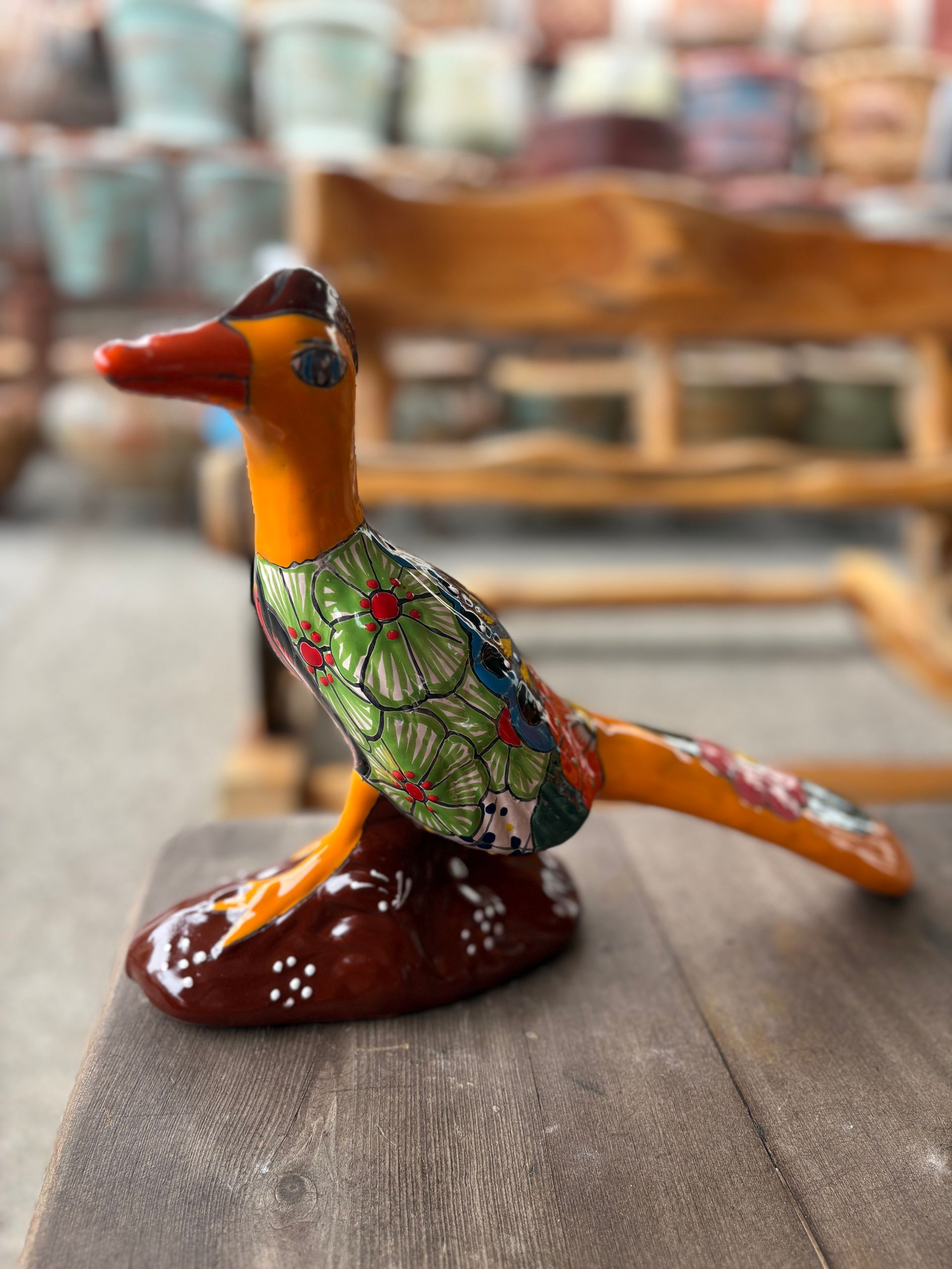 Ceramic roadrunner figurine with orange, green, and brown details, perched on a wooden surface in a shop.