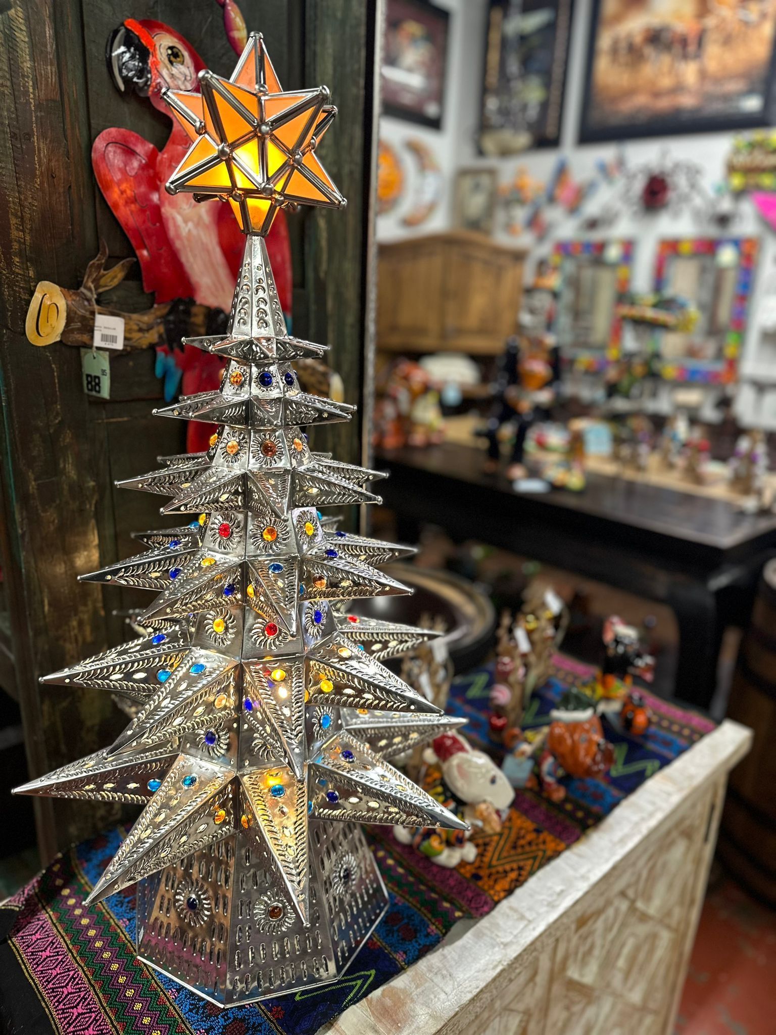 Silver metal Christmas tree-shaped lamp with colorful gems. The lamp is on a table in a shop filled with other items.