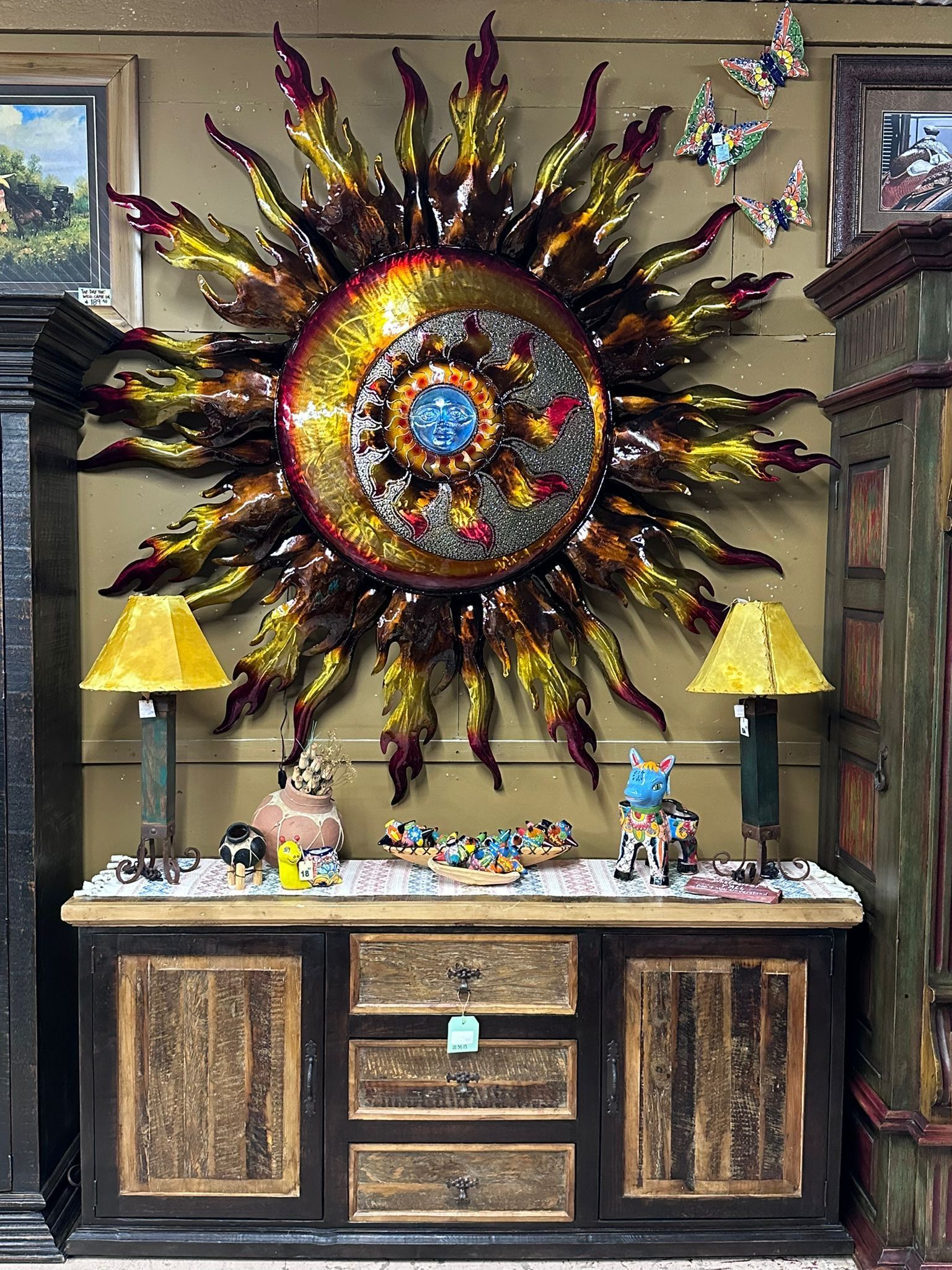 A display in a store featuring a large, colorful sunburst sculpture above a wooden cabinet. Two lamps flank the cabinet, with small decorative items on top.