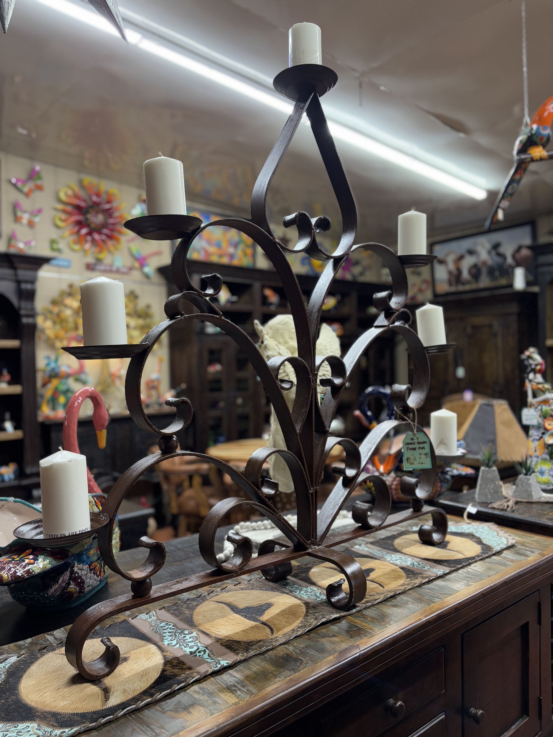 A wrought iron candelabra with nine white candles sits on a table. The candelabra has a decorative, swirled design and is in a store setting.