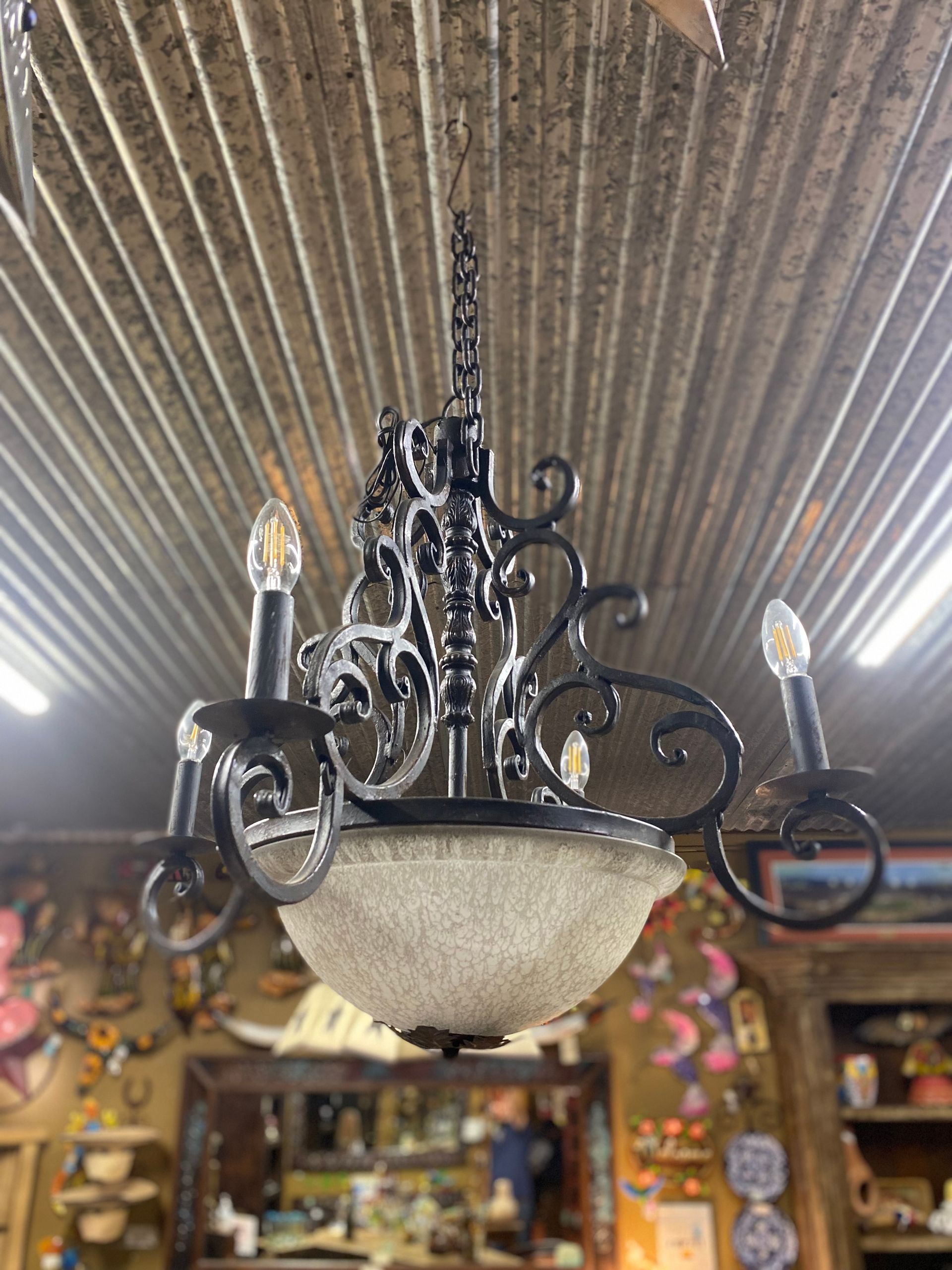 Ornate black chandelier with frosted glass bowl, hanging from a chain, against a corrugated metal ceiling.