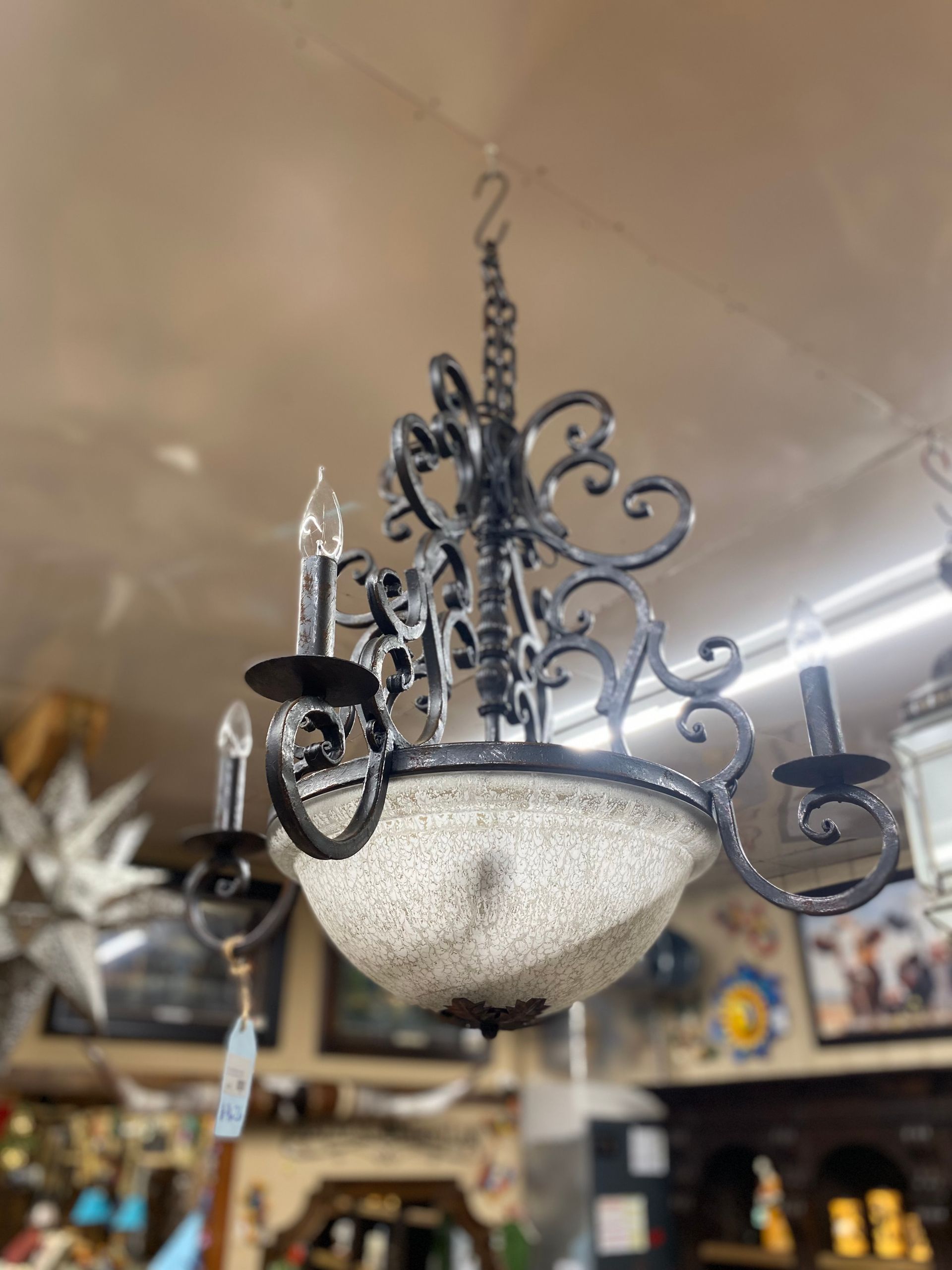 Black wrought iron chandelier with three candle-shaped lights and a speckled glass shade, hanging from a chain in a shop.