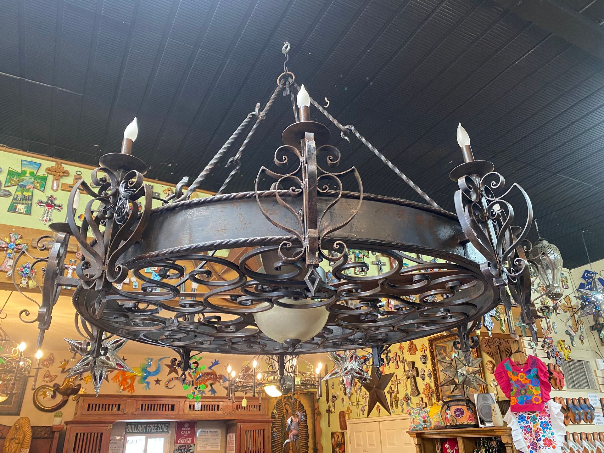 A large, ornate wrought iron chandelier hangs in a room with a black ceiling. Decorative wall art surrounds the chandelier.