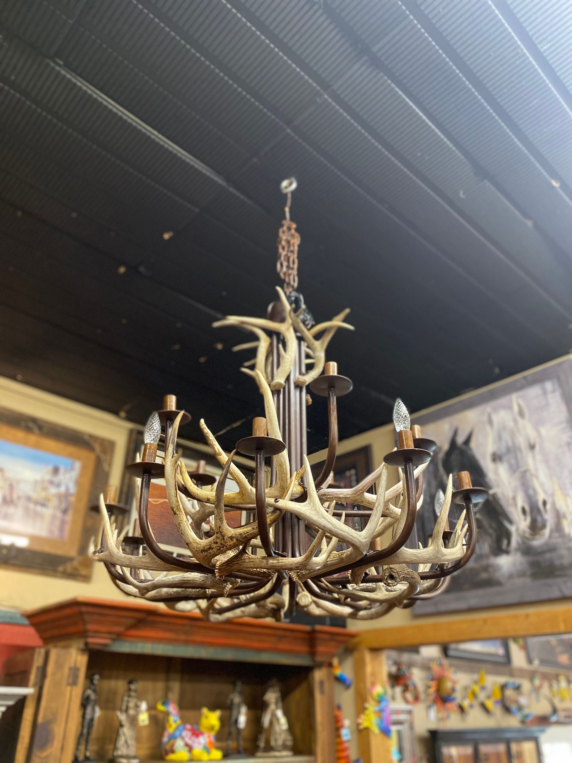 A rustic antler chandelier hangs from a dark ceiling. The fixture features multiple light bulbs and is in a room with artwork.
