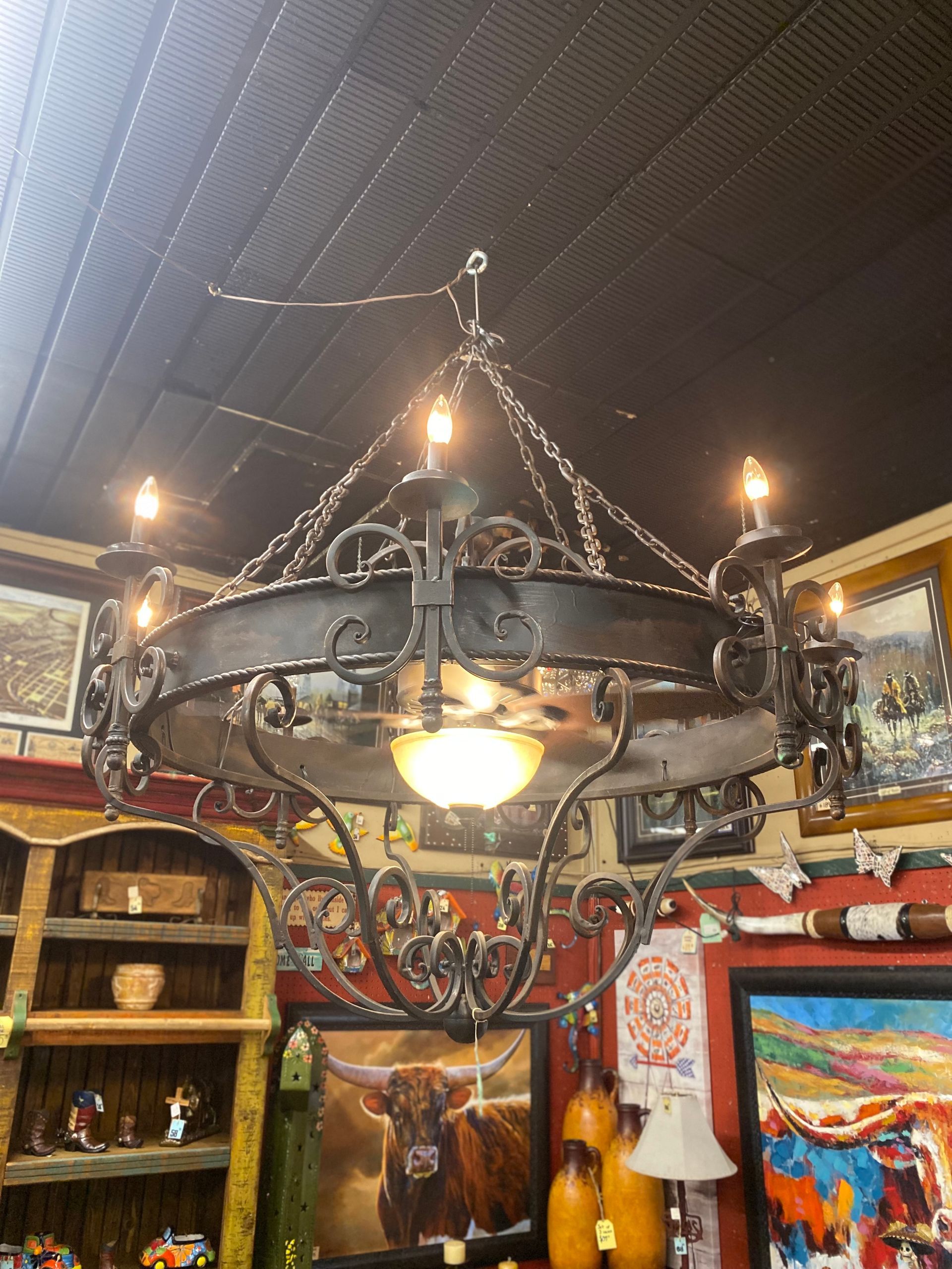 Rustic metal chandelier with six candle-style lights, suspended from a dark ceiling. The fixture is in a shop filled with artwork and decor.
