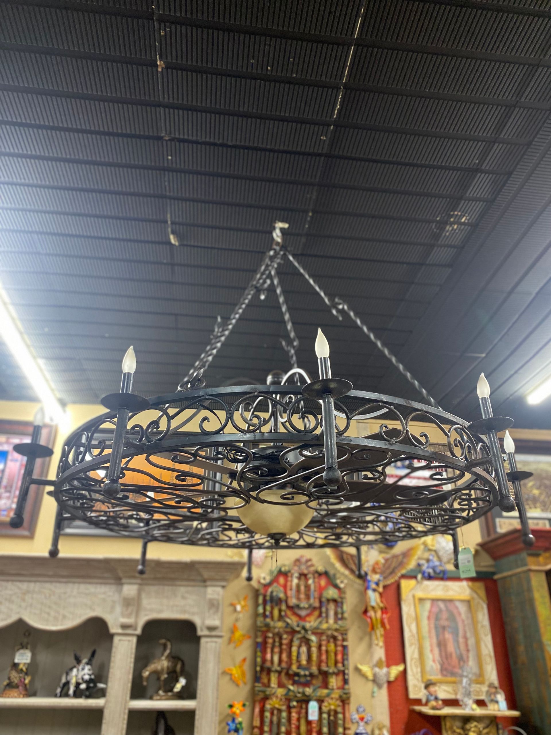 Ornate wrought iron chandelier hanging from a black ceiling in a store. It has several light bulbs and decorative flourishes.