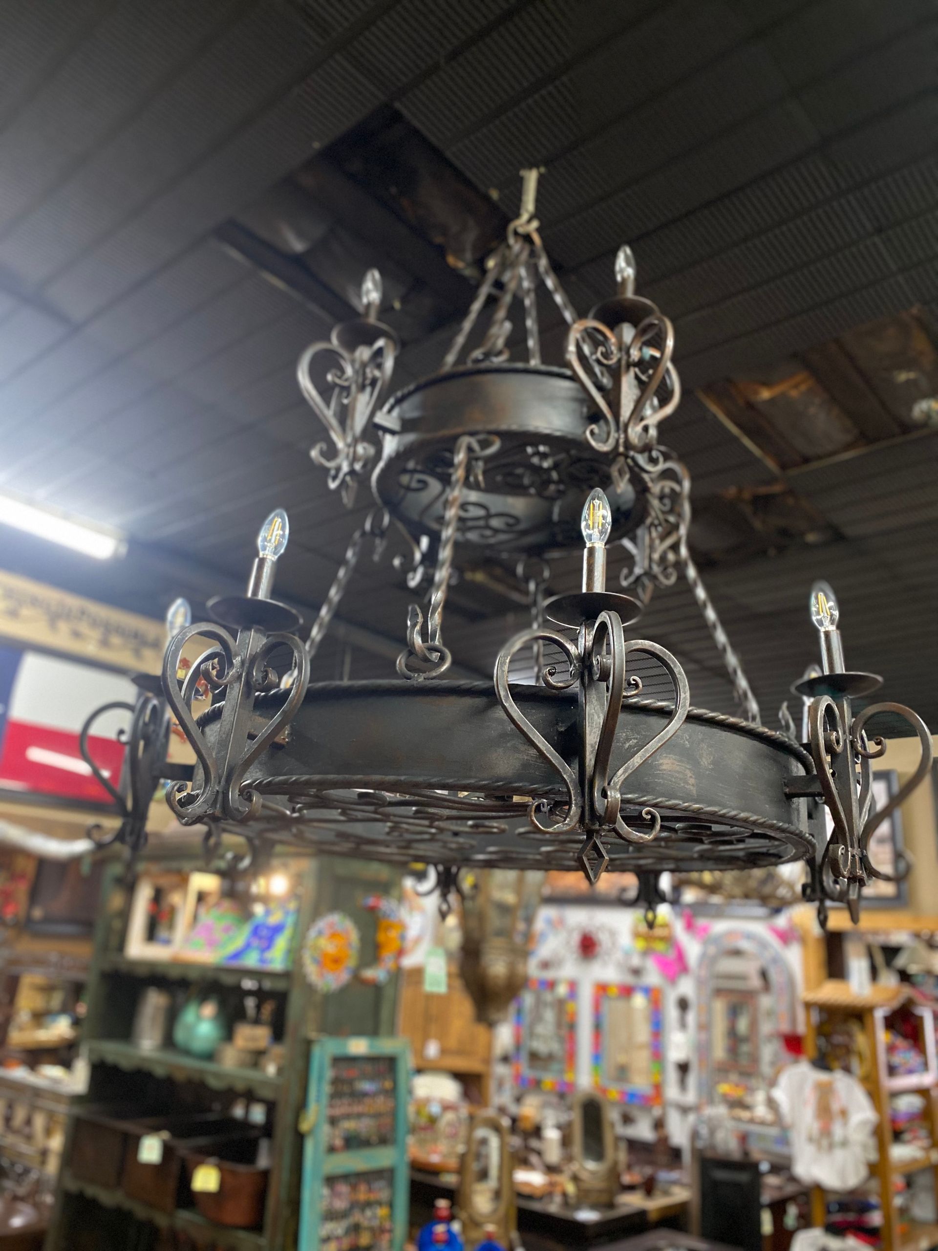Black wrought iron chandelier with ornate scrollwork hanging in a cluttered antique shop.